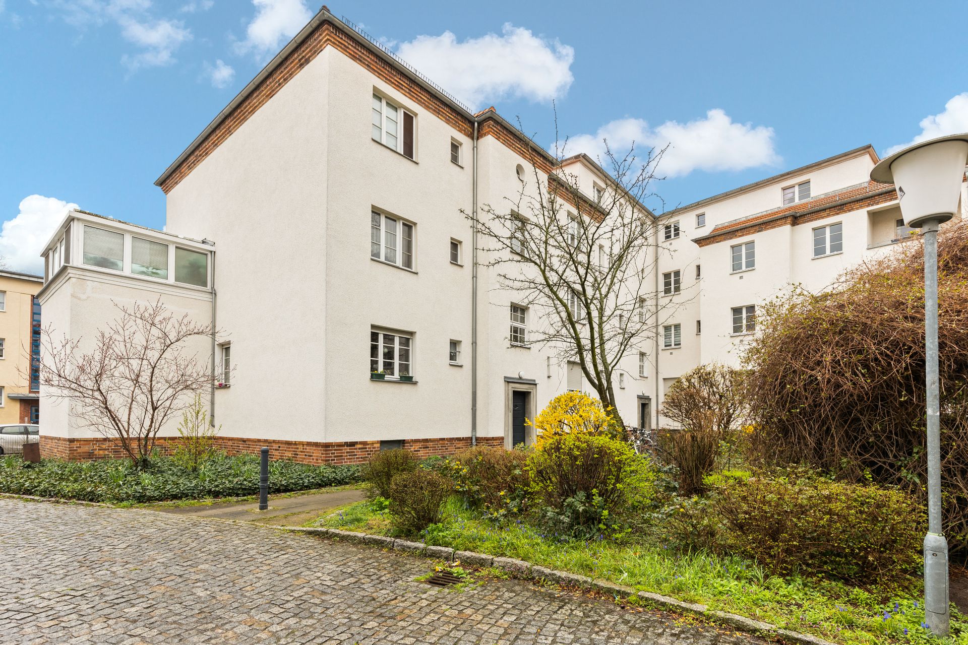Mehrstöckiges Wohngebäude mit hellem Putz, Vorgarten und Baum, gepflasterter Weg im Vordergrund.