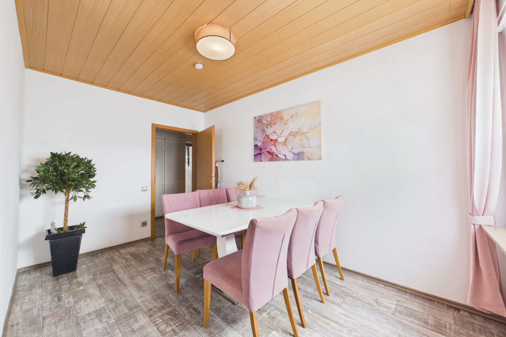 Esszimmer mit weißem Tisch, vier rosa Stühlen, Pflanze und großem Fenster mit rosa Vorhängen.