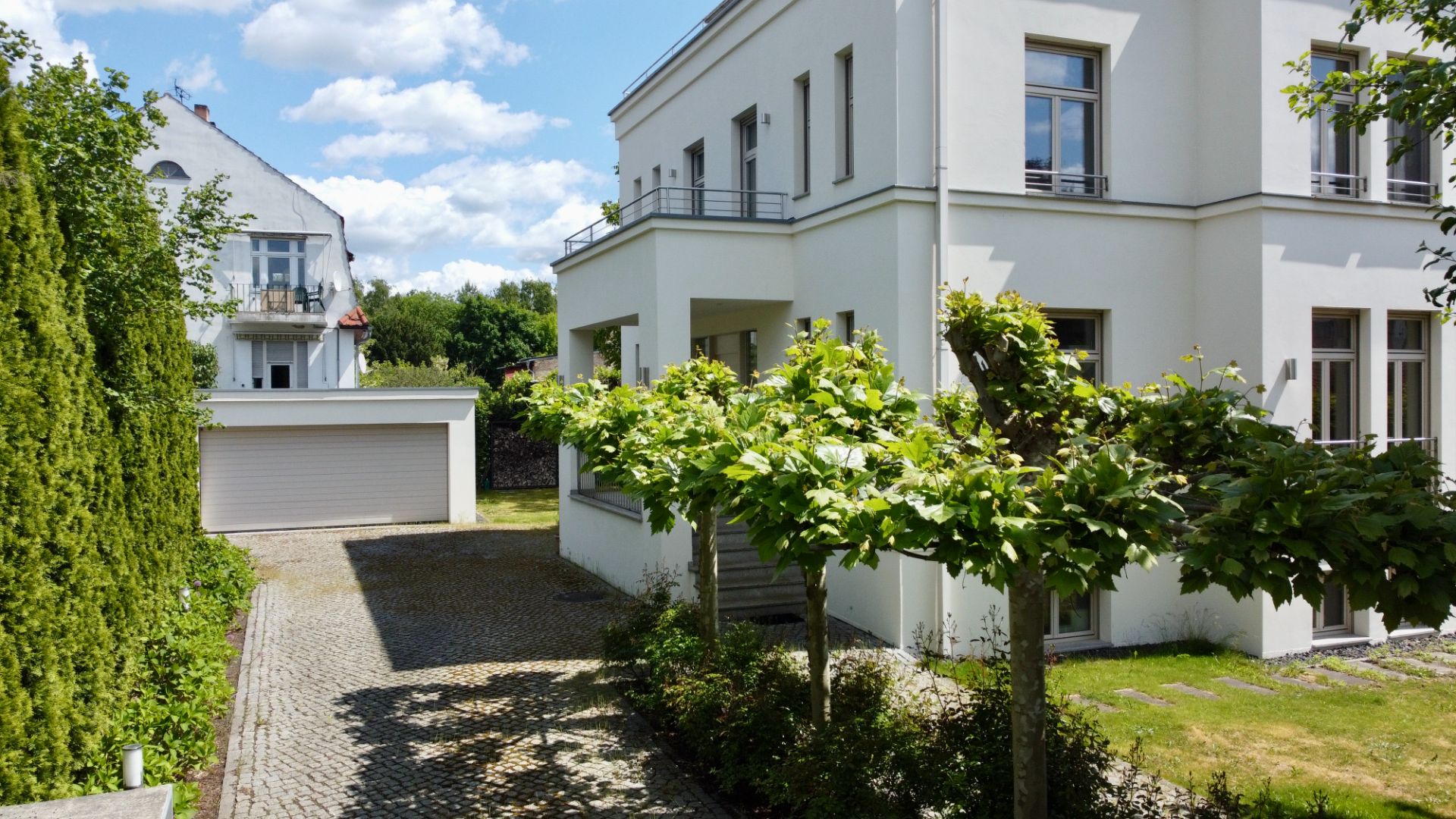 Moderner weißer Wohnhausbau mit Bäumen im Garten und einer gepflasterten Einfahrt zu einer Garage.