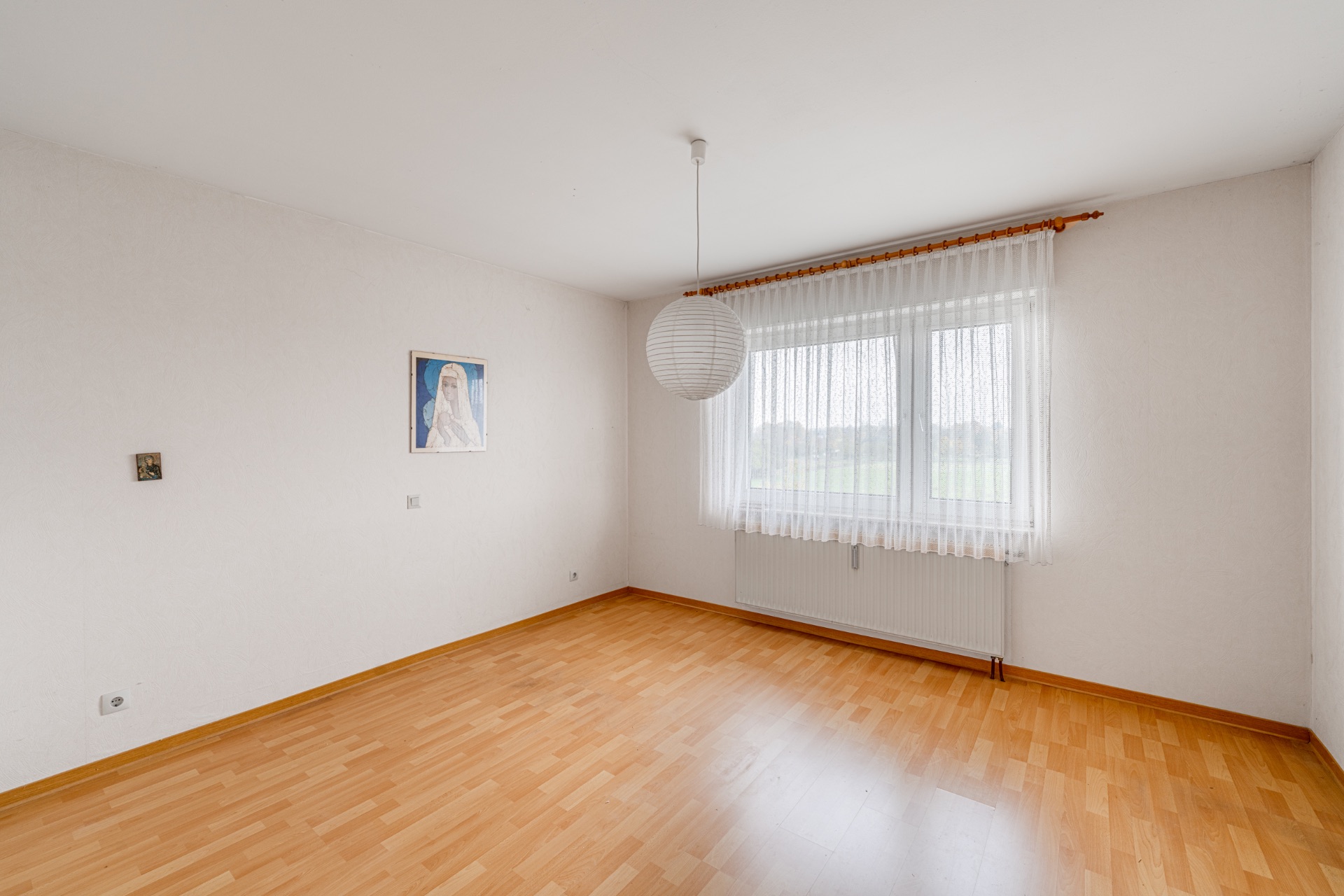 Leeres Zimmer mit Holzfußboden, großem Fenster mit Vorhang und zwei Bildern an der Wand.