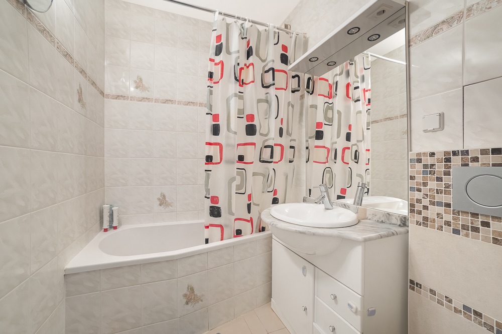 Helles Badezimmer mit Badewanne, gemusterter Duschvorhang und Waschbecken mit Spiegel.