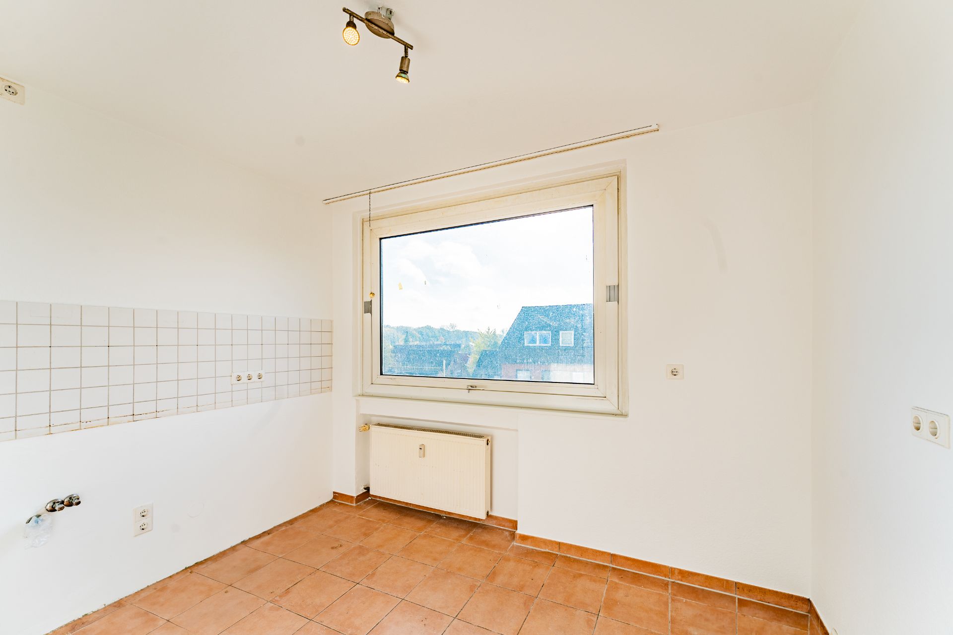 Leerer Raum mit Fliesenboden, weißer Wand, großem Fenster und Heizkörper.