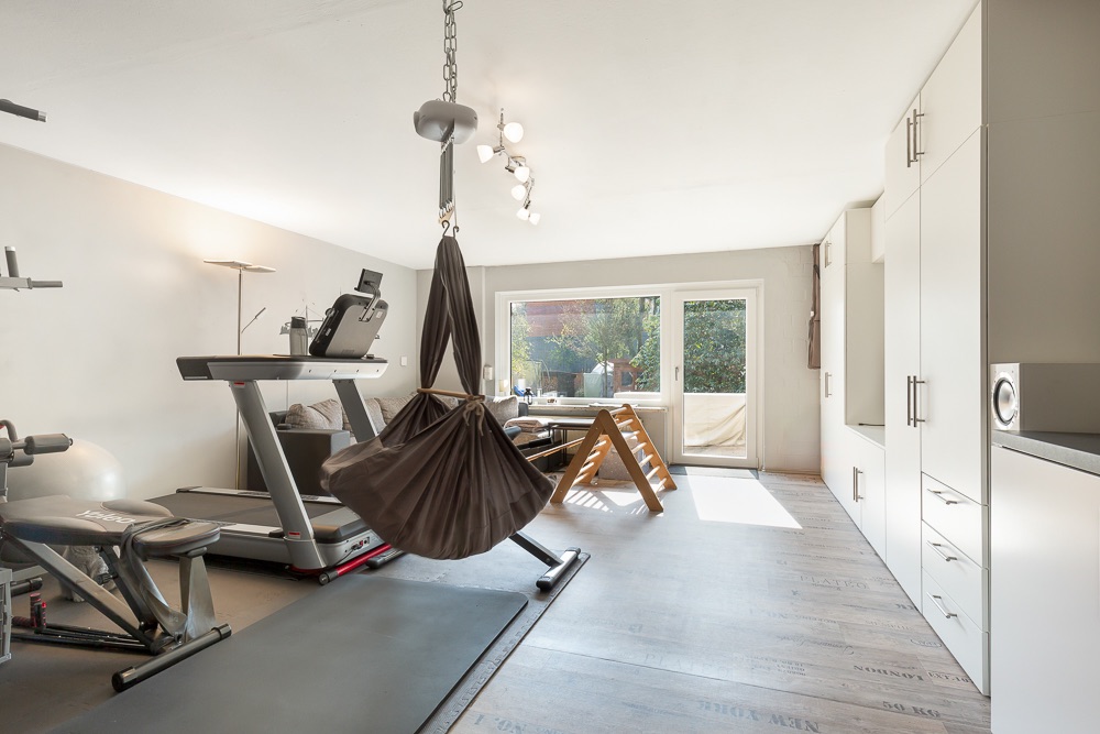 Heller Raum mit Fitnessgeräten, Hängesessel und großen Fenstern zu einem Garten.