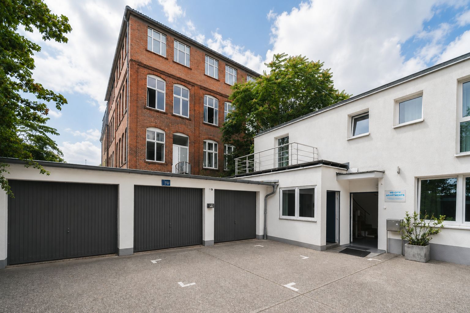 Hof mit drei Garagentoren, weißem Wohngebäude und rotem Backsteinhaus im Hintergrund.