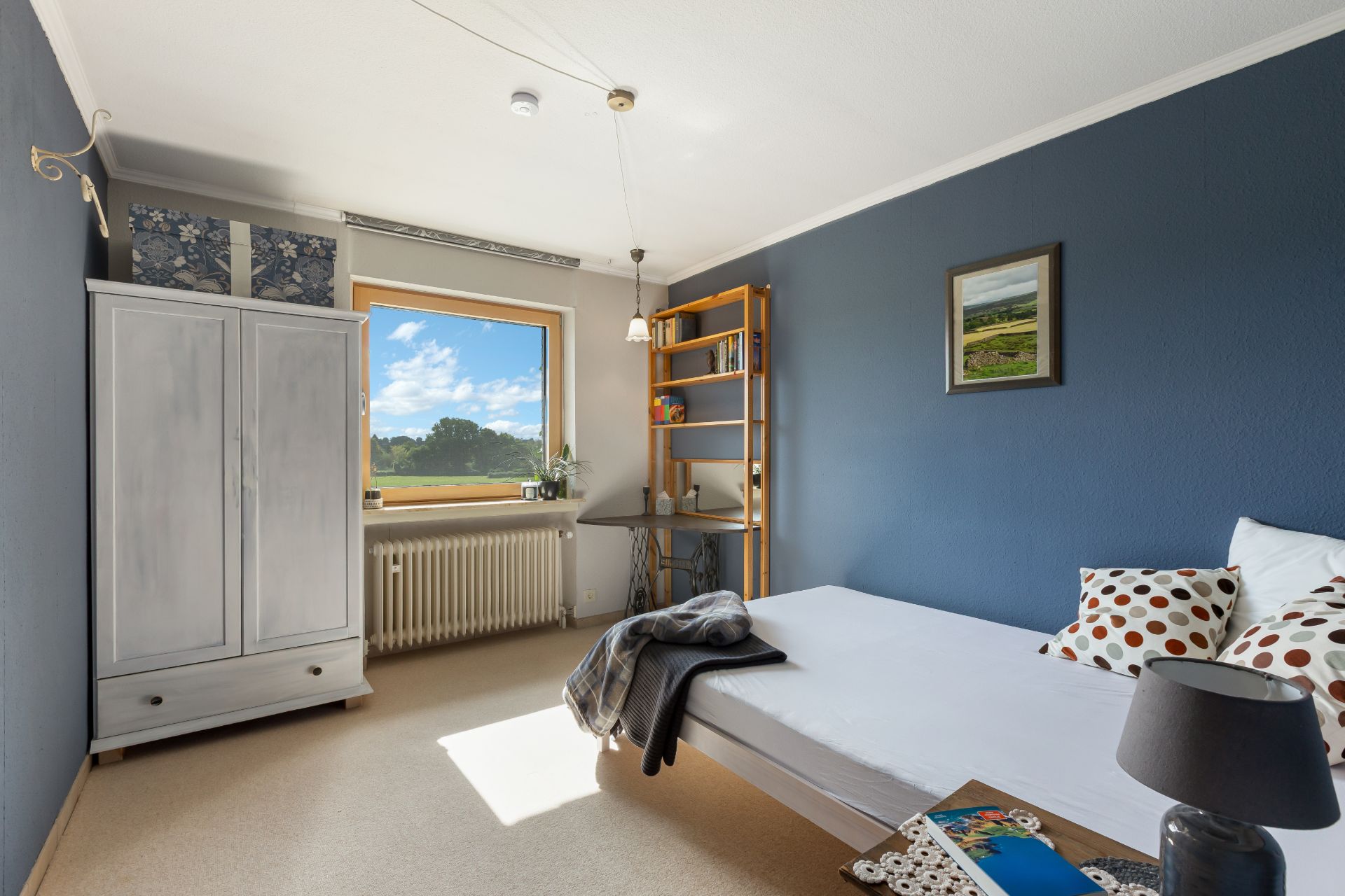 Helles Schlafzimmer mit Bett, weißem Schrank, Regal, Fenster und blau gestrichener Wand.