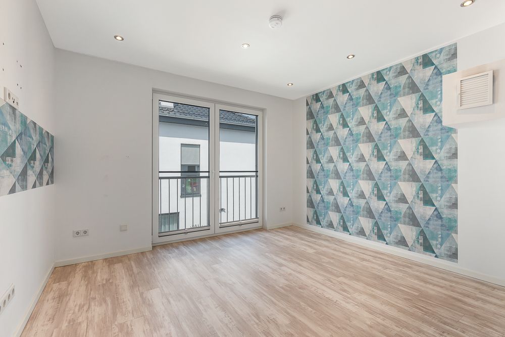 Helles Zimmer mit Holzboden, Musterwand und großer Glastür zum Balkon.