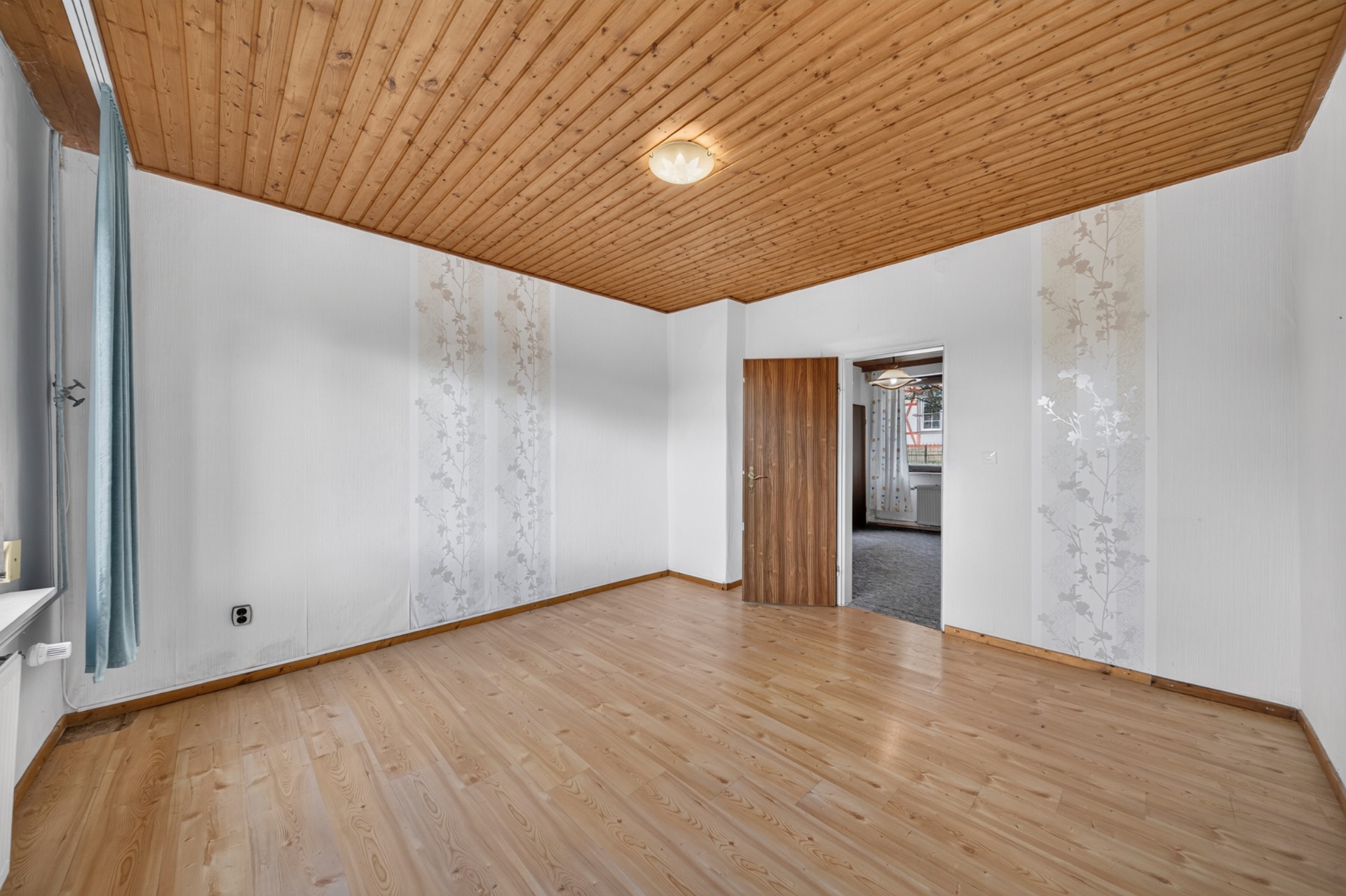 Leerer Raum mit Holzdecke, hellem Boden und weiß gestrichenen Wänden, eine Tür führt in einen anderen Raum.