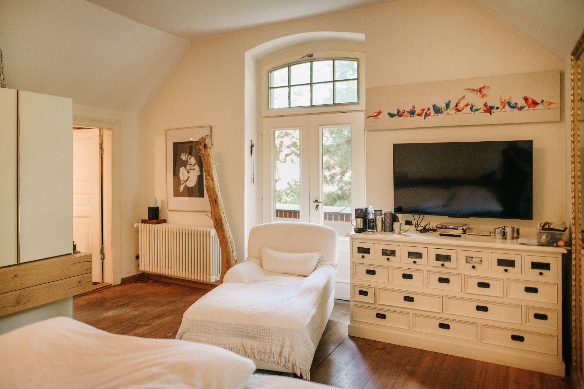 Helles Schlafzimmer mit Sessel, Kommode, großem Fernseher und Fenster mit Blick nach draußen.