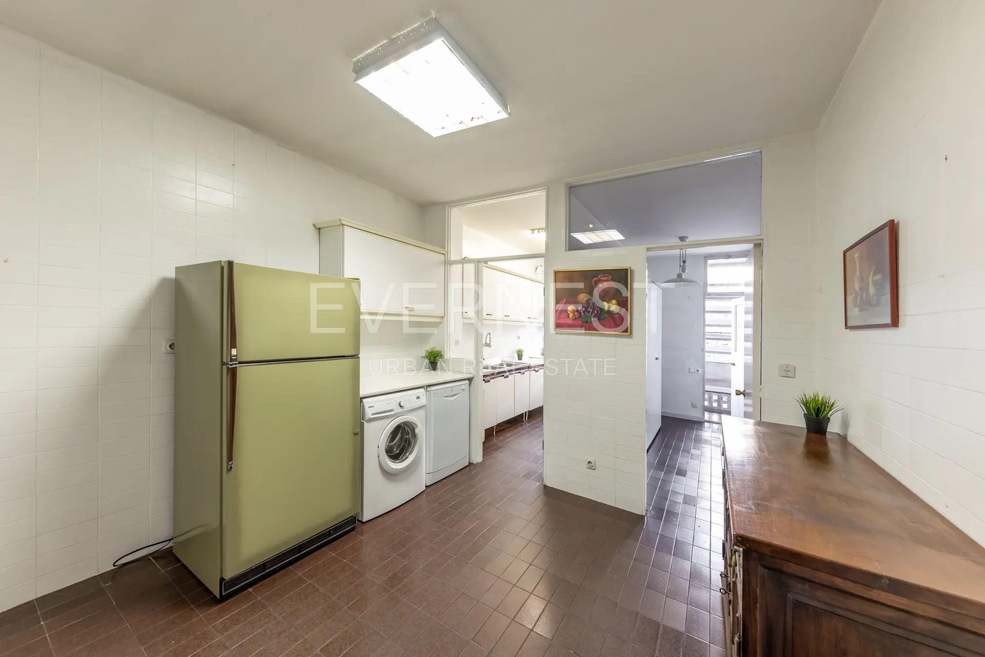 Helle Küche mit großem Kühlschrank, Waschmaschine und weißen Wänden, brauner Fliesenboden.