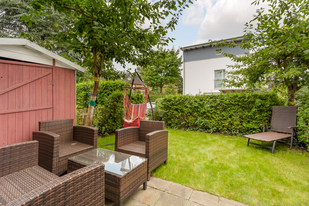 Garten mit Sitzgruppe aus Rattan, Liege, Schaukelstuhl und rotem Holzschuppen.