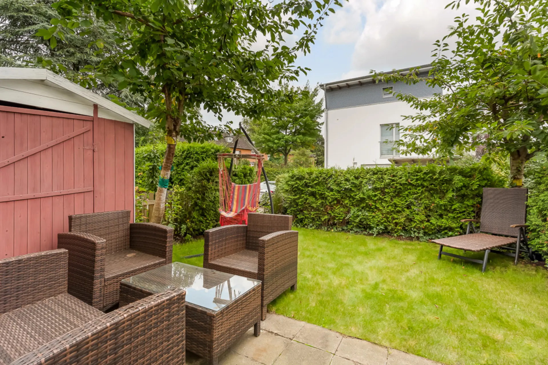 Garten mit Sitzgruppe aus Rattan, Liege, Schaukelstuhl und rotem Holzschuppen.