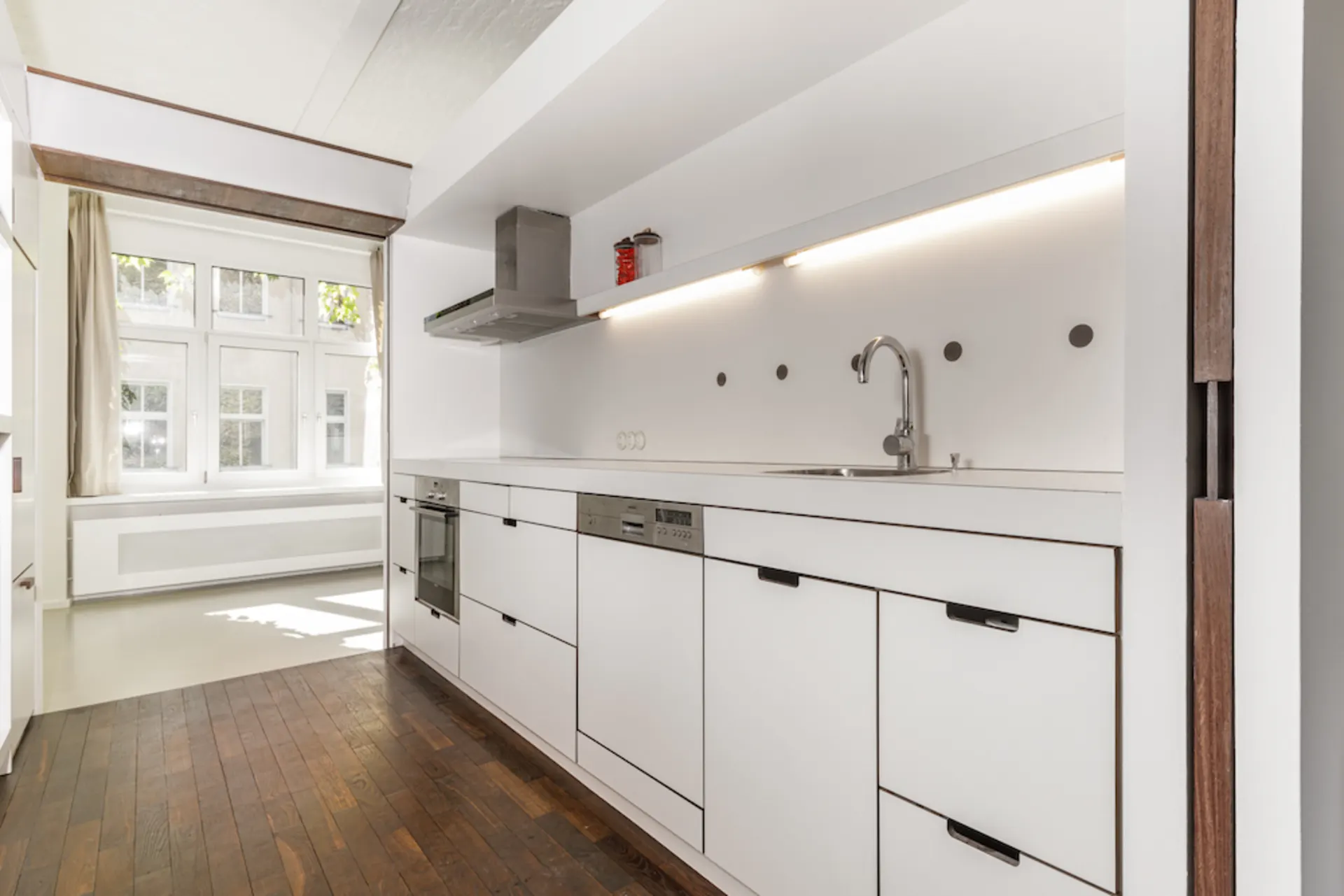 Moderne weiße Küche mit Spüle, Ofen und großem Fenster, heller Holzboden.
