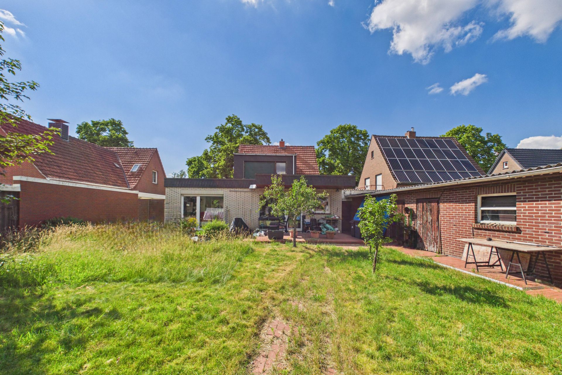 Garten mit Rasen und Wegen, im Hintergrund ein Haus mit Terrasse, Bäumen und Nachbarhäusern.