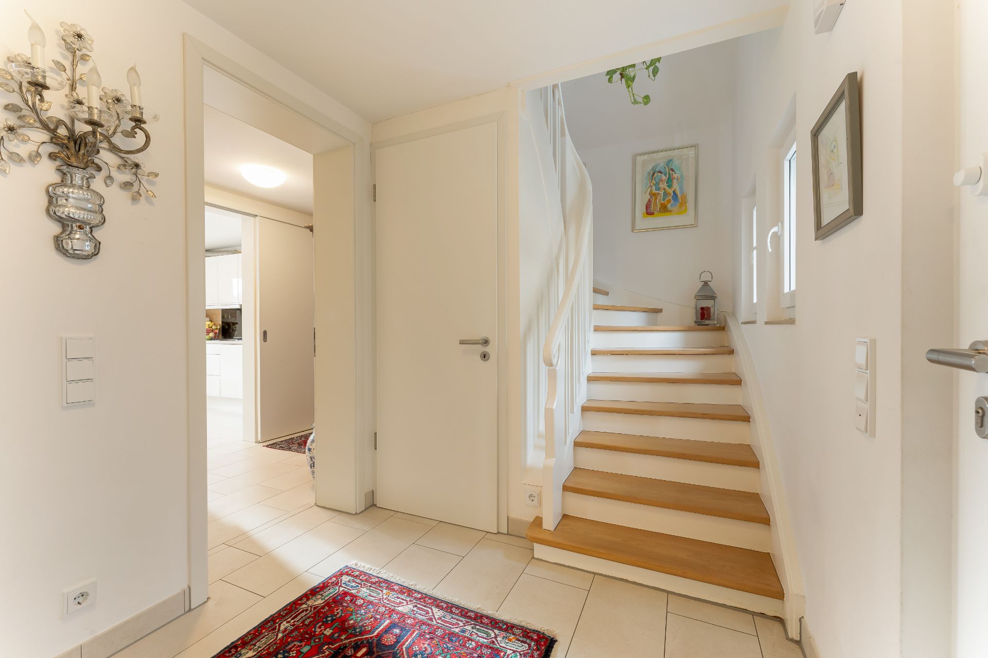 Heller Flur mit Treppe, Teppich und Wandbildern, Durchgang zur Küche sichtbar.