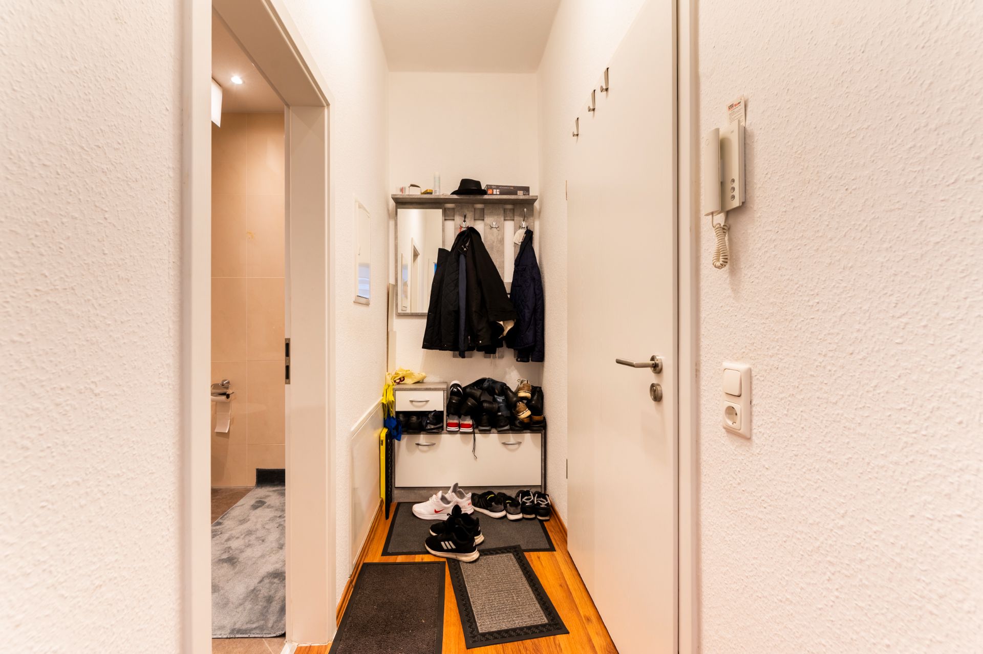 Flur mit Garderobe, mehreren Jacken, Schuhen, Teppichen und einem Blick ins Badezimmer.