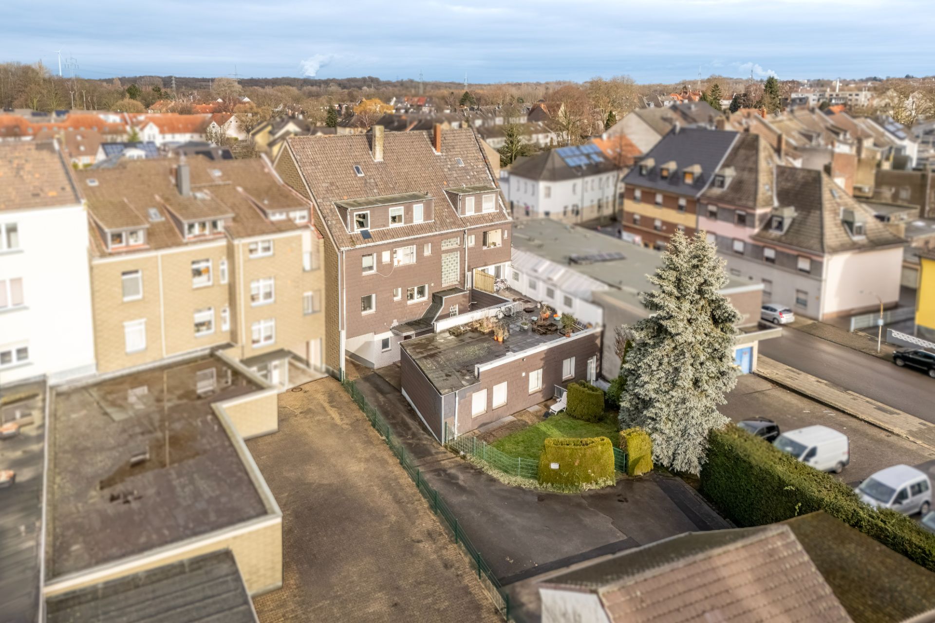Mehrfamilienhaus mit flachem Anbau, Baum und kleinem Garten, umgeben von weiteren Wohnhäusern.