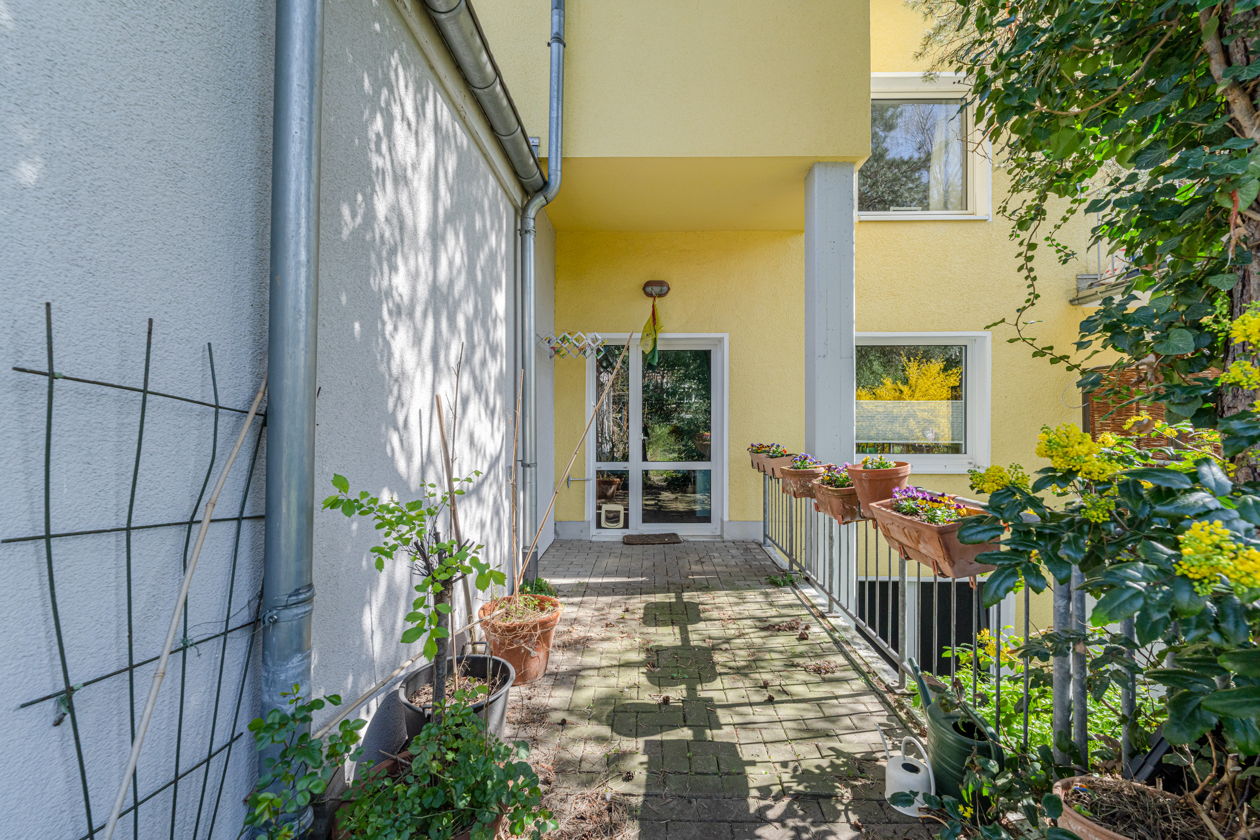Eingangsbereich eines Wohnhauses mit gelber Fassade, Pflanzenkübeln und Blumenkästen am Geländer.