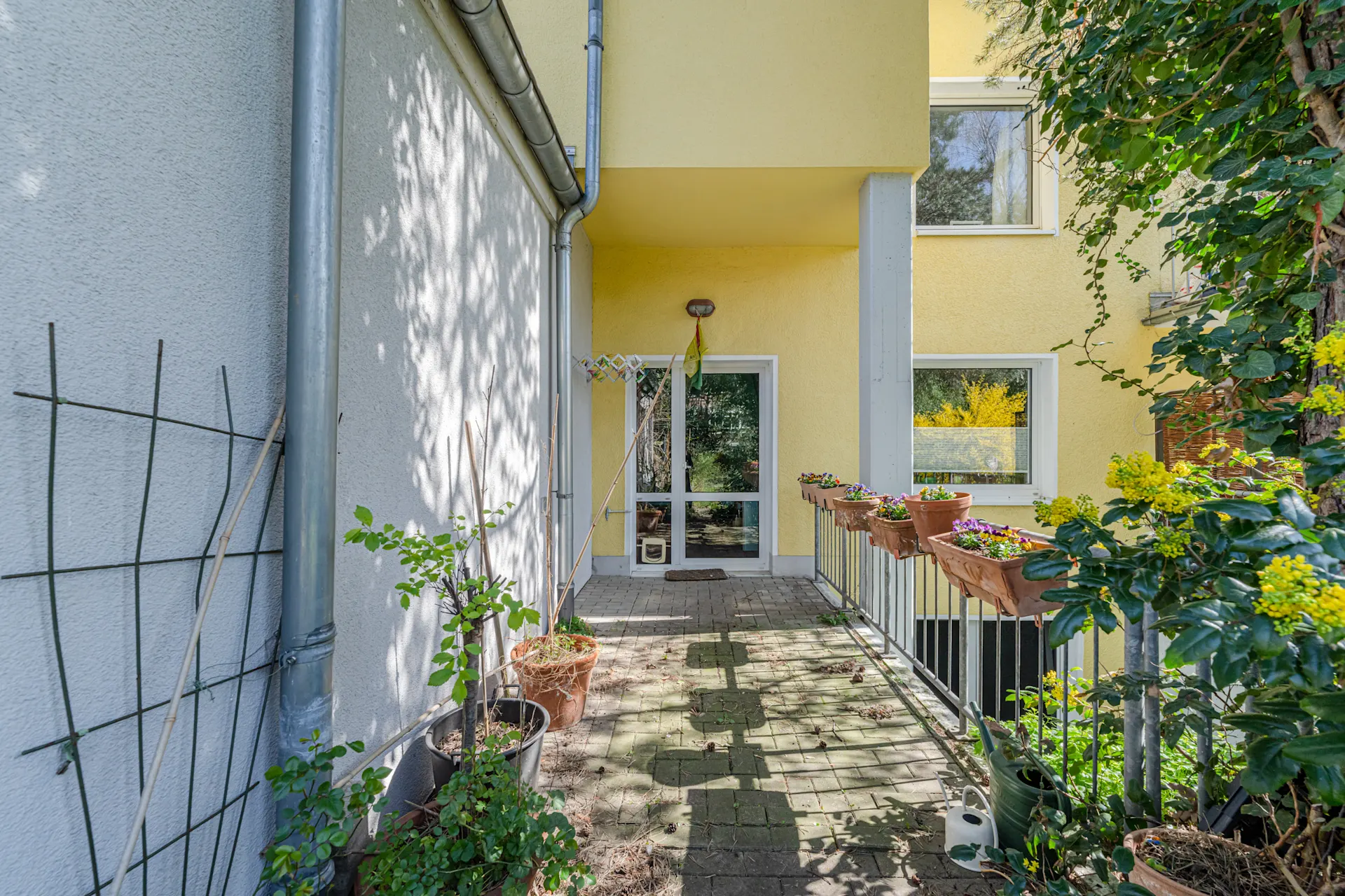 Eingangsbereich eines Wohnhauses mit gelber Fassade, Pflanzenkübeln und Blumenkästen am Geländer.