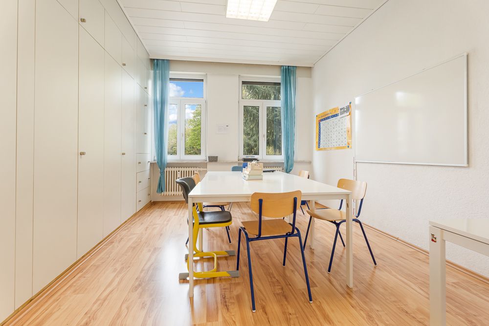 Heller Raum mit Tisch und Stühlen, großen Schränken, zwei Fenstern und Whiteboard an der Wand.