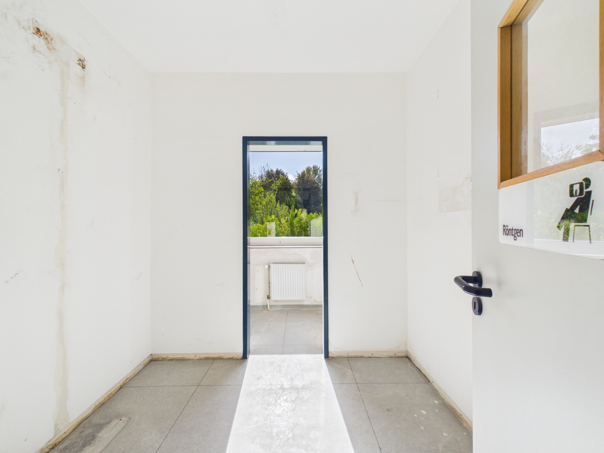 Heller Raum mit weißen Wänden, offenem Türrahmen und Blick auf einen Balkon mit Grün im Hintergrund.