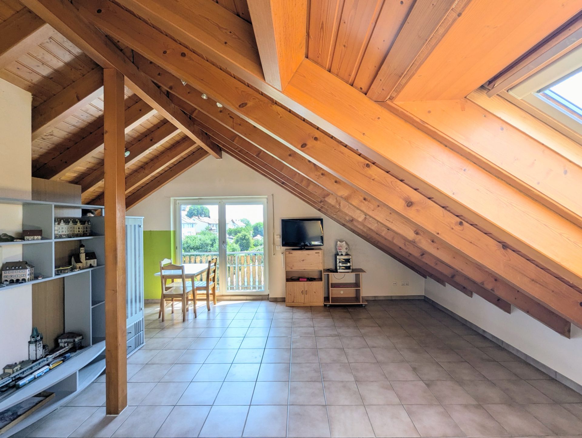 Heller Dachbodenraum mit Holzbalken, Fliesenboden, Regal, Fernseher und Esstisch am Fenster.