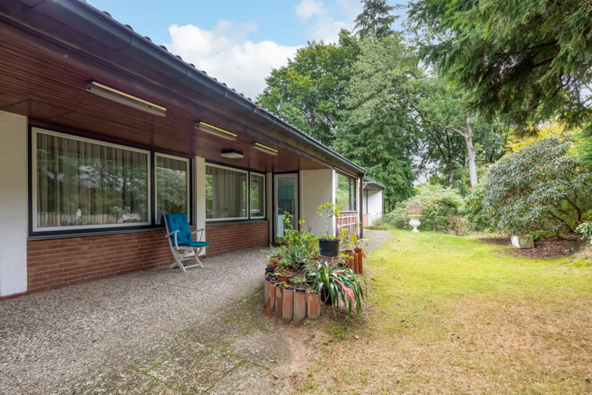 Überdachte Terrasse mit Stuhl und Pflanzgefäßen, angrenzend an einen Garten mit Bäumen und Sträuchern.