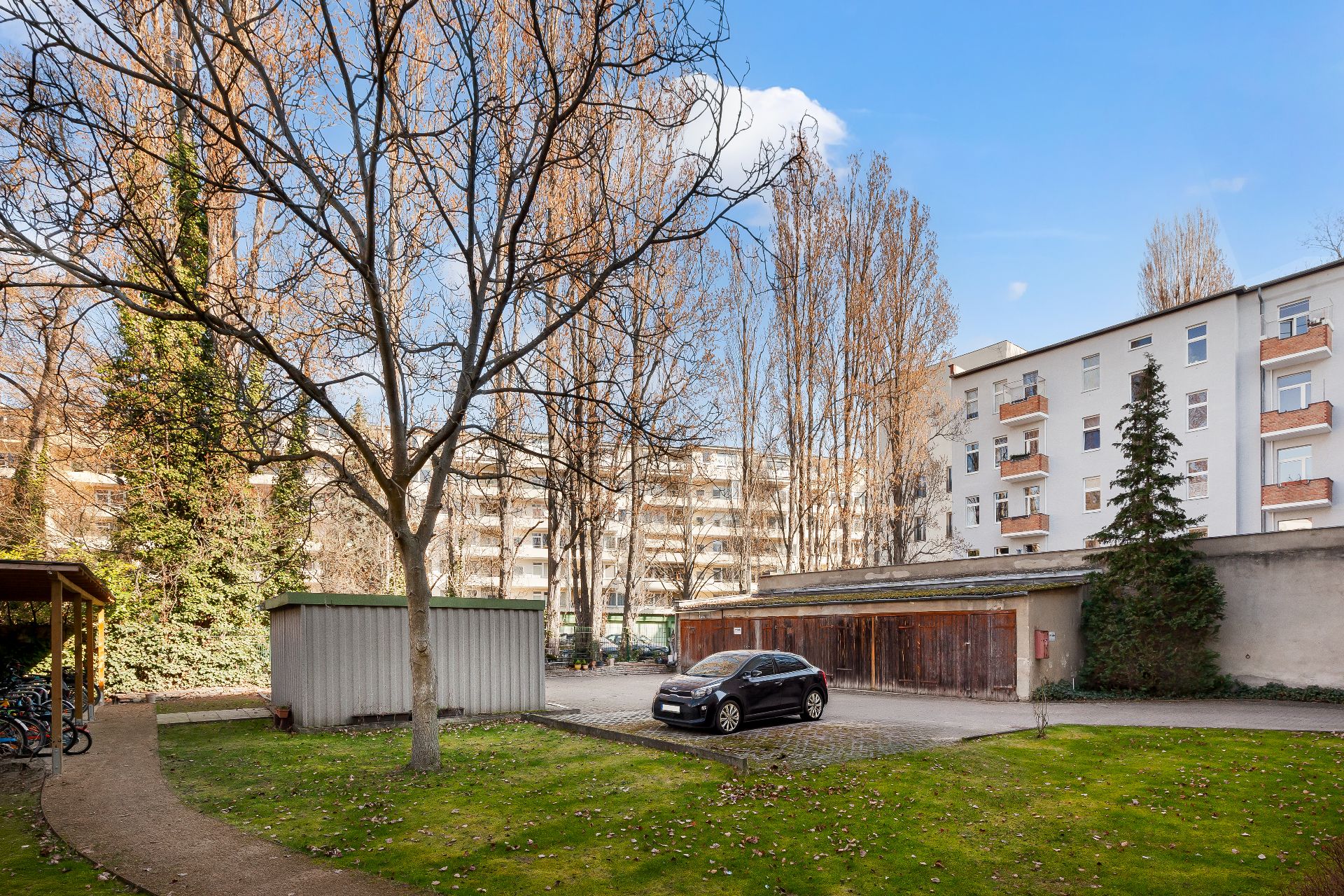 Grüner Innenhof mit Bäumen, einem Auto auf einem Parkplatz und Wohnhäusern im Hintergrund.