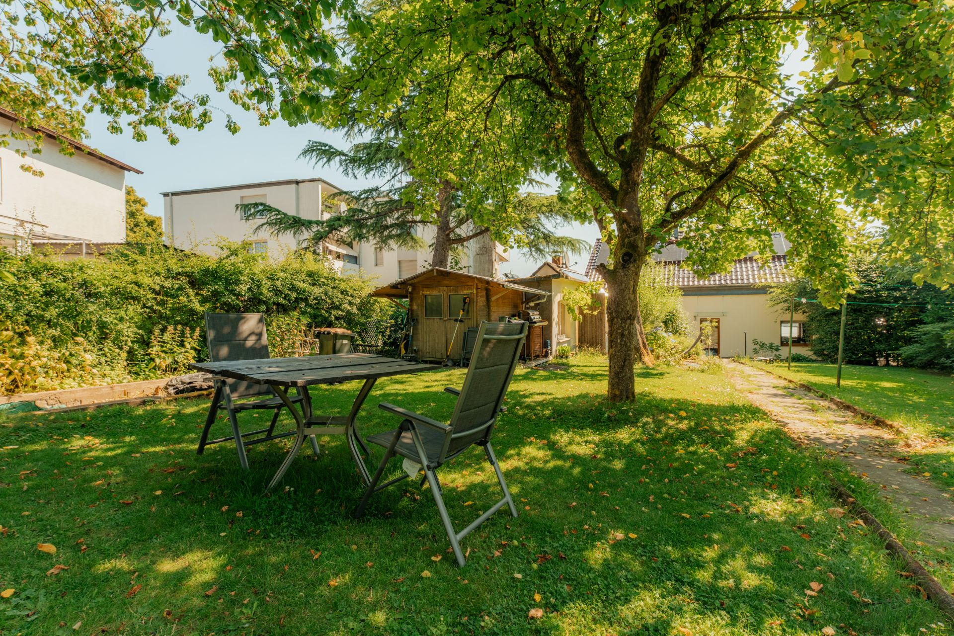 Grüner Garten mit Tisch, zwei Stühlen, Baum und kleinem Holzhaus im Hintergrund.
