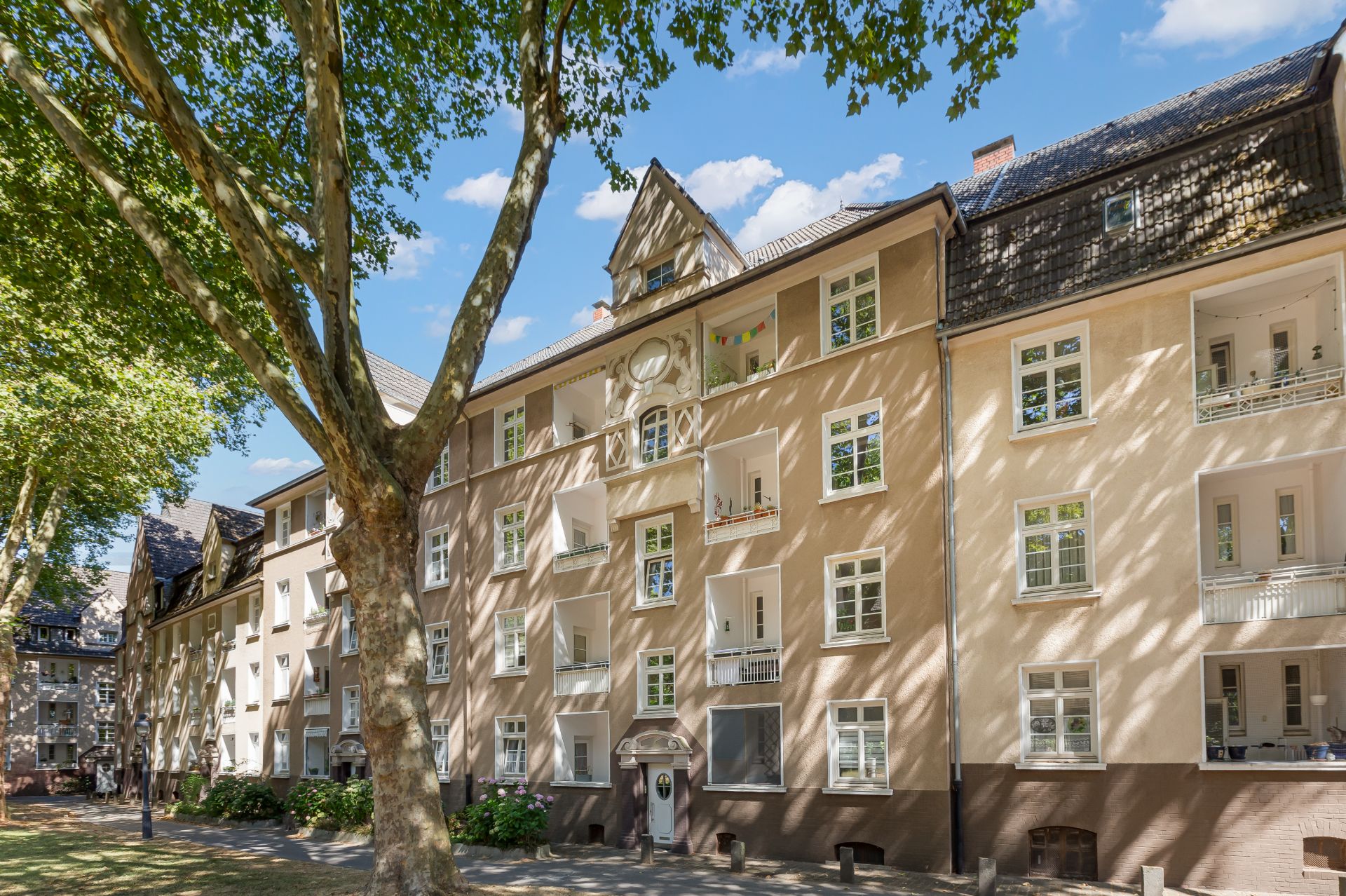 Mehrstöckiges Wohngebäude mit mehreren Balkonen und großen Fenstern, davor Bäume und Rasen.