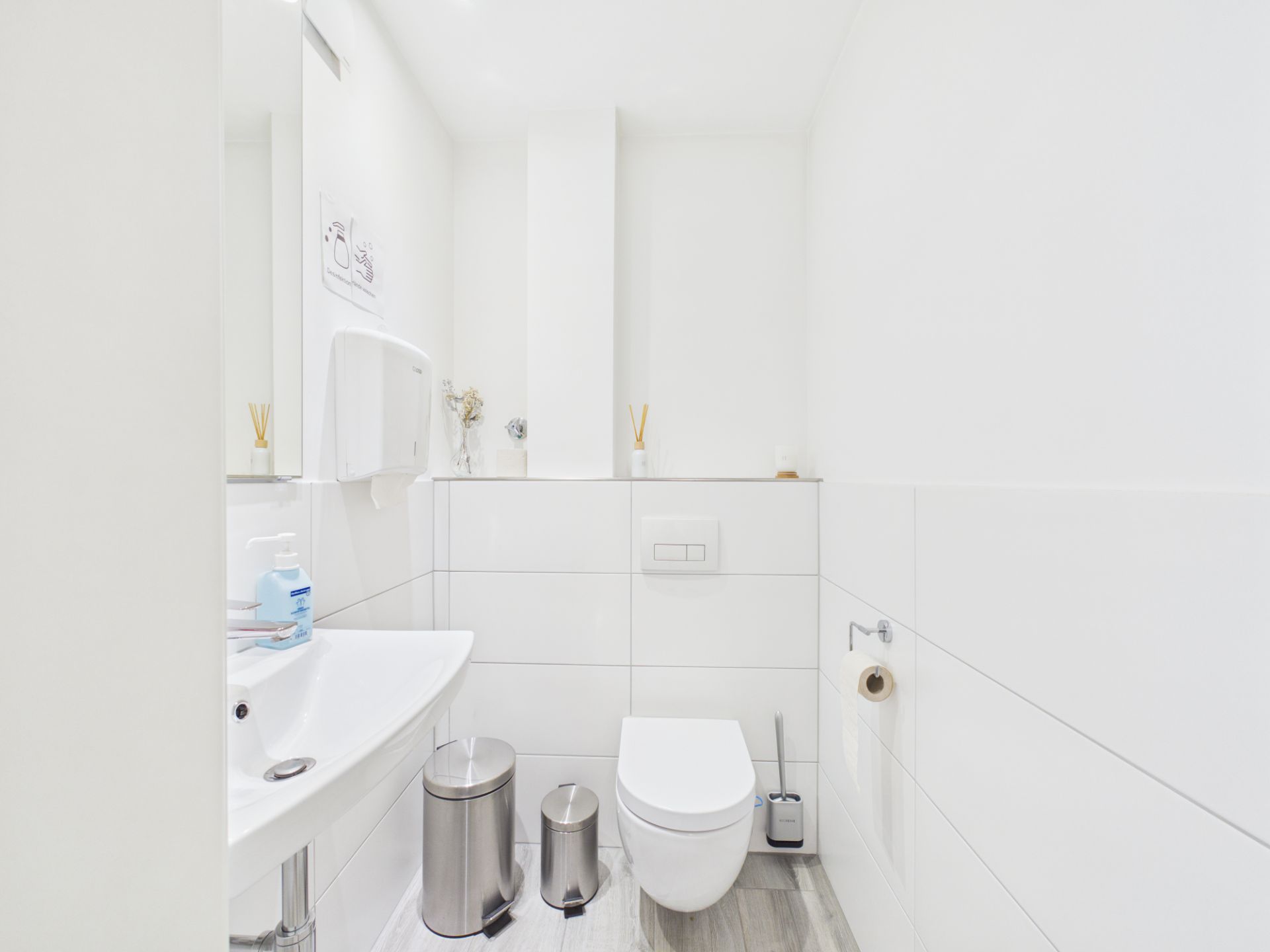 Modernes, weiß gefliestes Badezimmer mit Wandtoilette, Waschbecken und zwei Mülleimern.