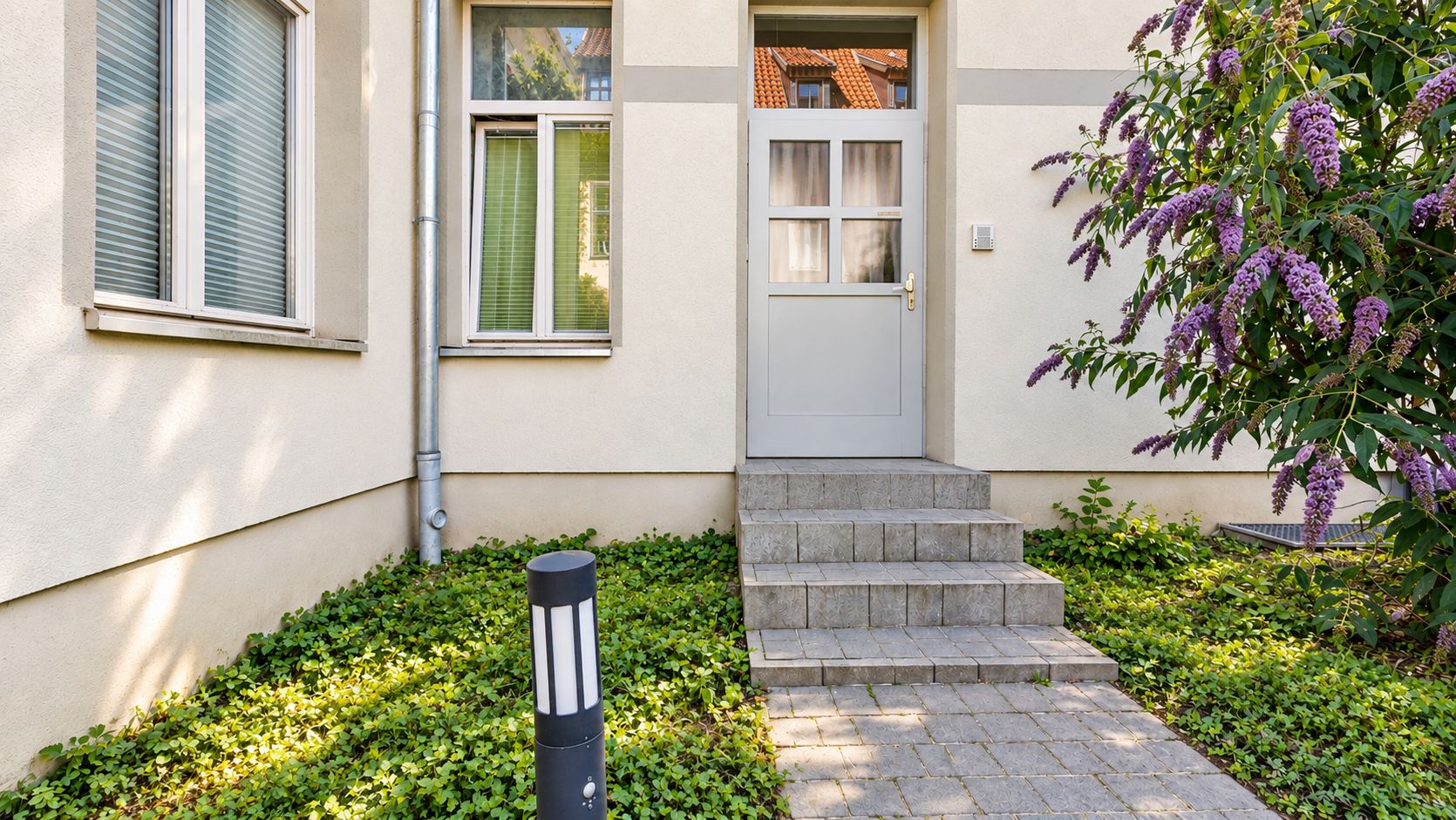 Eingangstür eines Wohnhauses mit Treppe, Pflasterweg, Beet und blühendem Busch rechts.