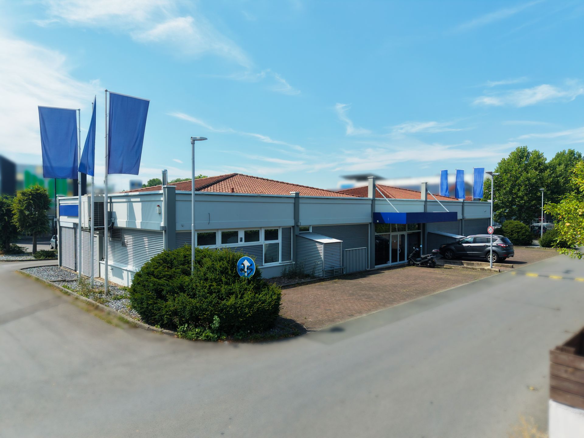 Flaches Gebäude mit rotem Dach, großen Fenstern und blauen Fahnen, Parkplatz davor.