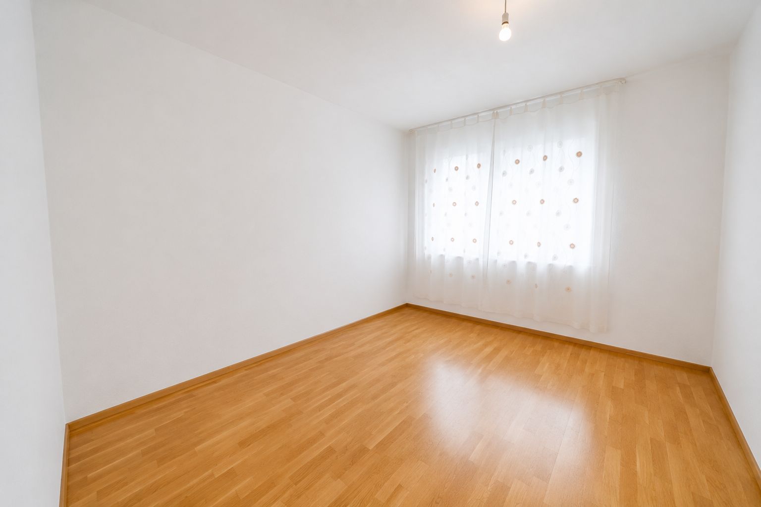 Leeres Zimmer mit Parkettboden, weißen Wänden und Fenster mit weißen Gardinen.