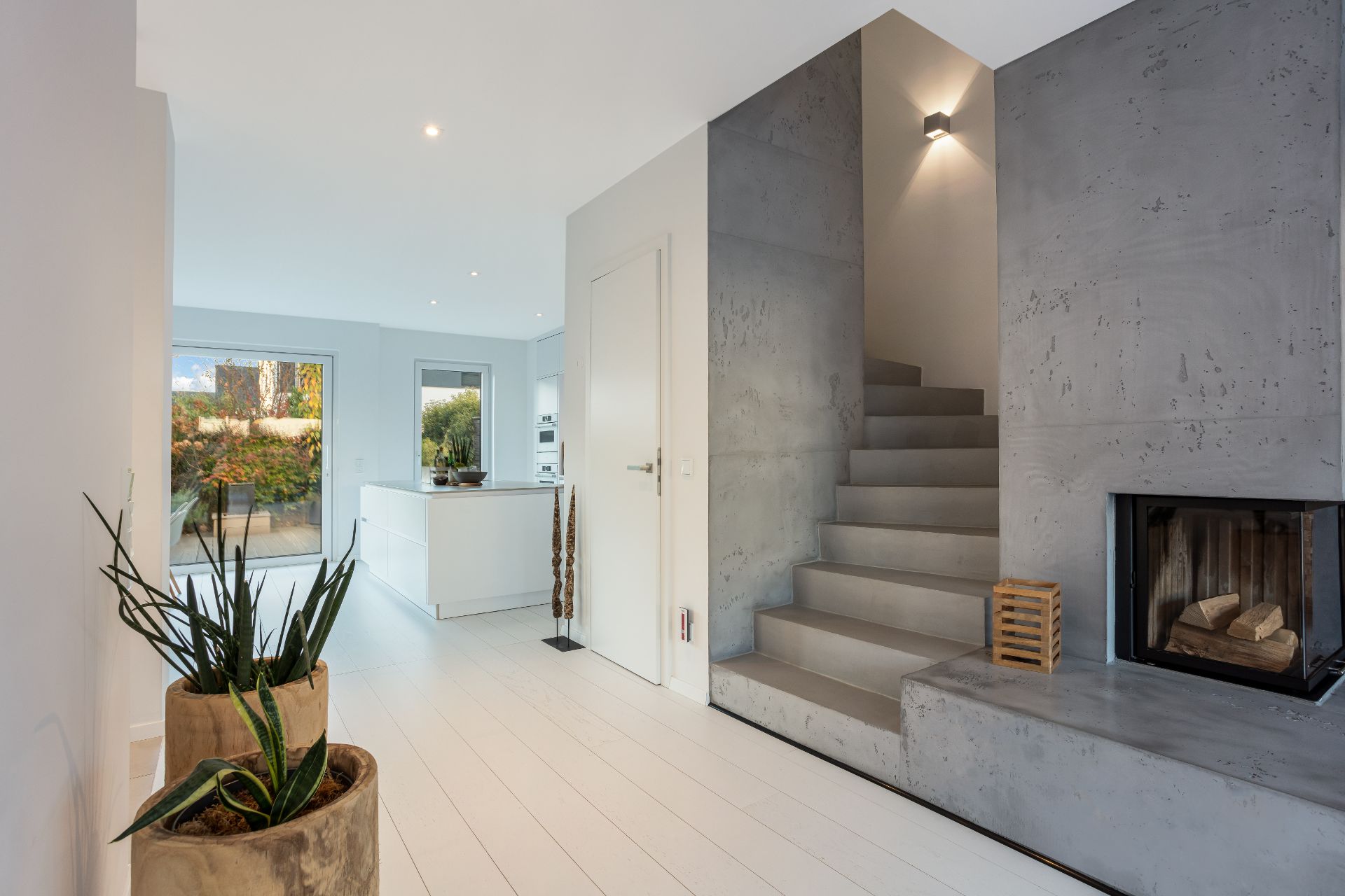 Moderner Wohnbereich mit Kamin, Treppe, Pflanzen und offener Küche mit Blick nach draußen.