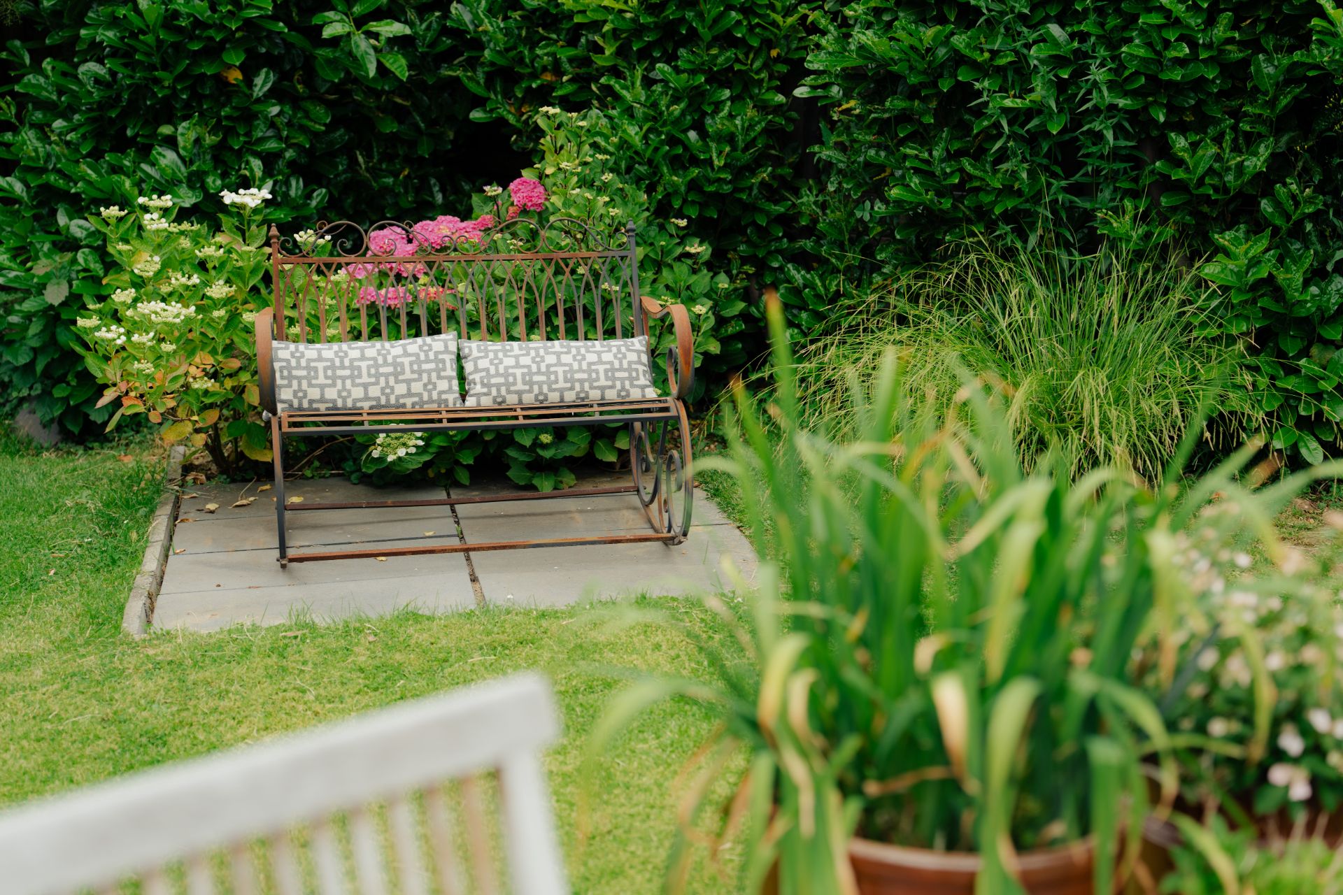 Metallbank mit Kissen im Garten, umgeben von Blumen und grünen Büschen.