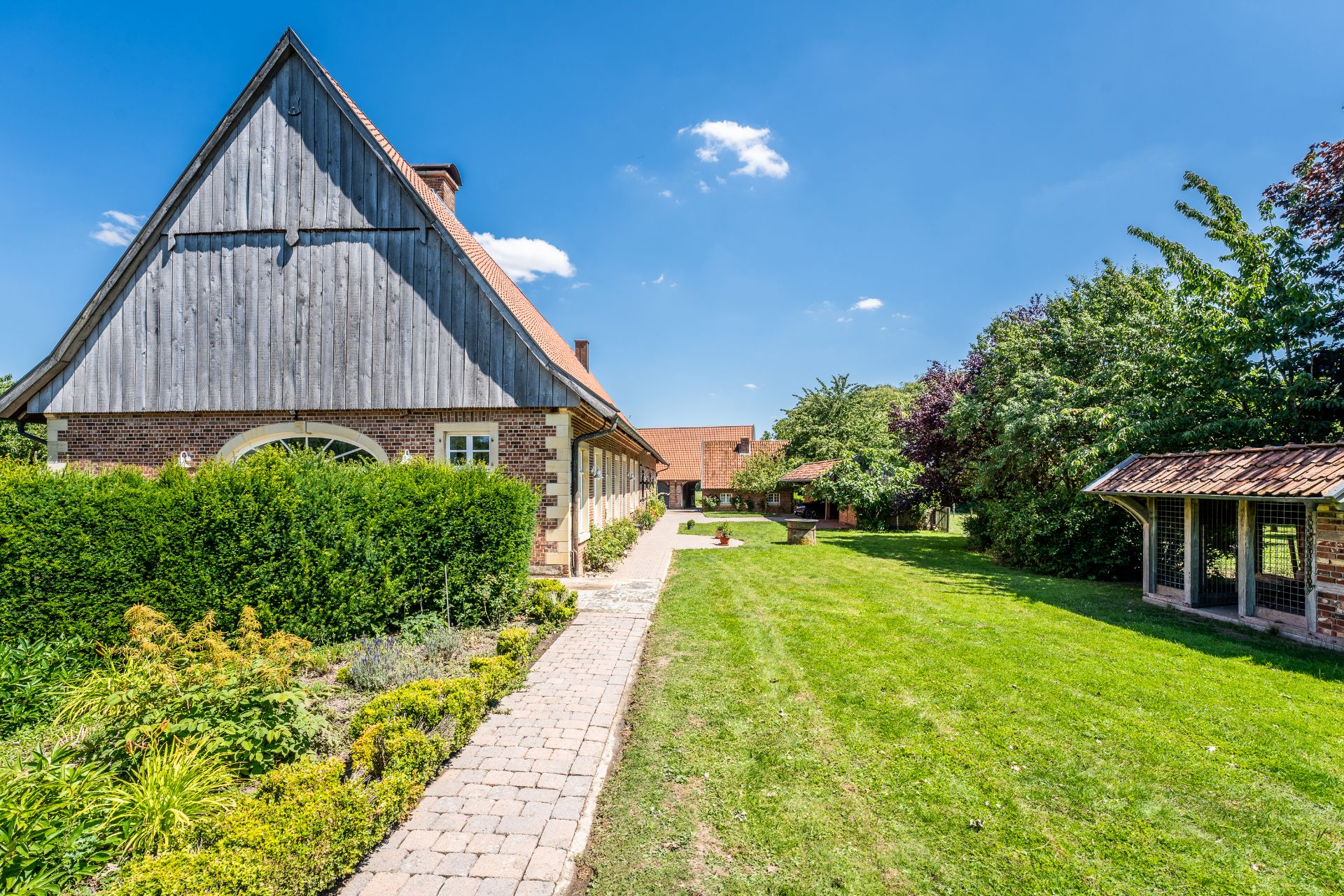Großes Haus mit Spitzdach, Garten, gepflastertem Weg und Nebengebäude, umgeben von Bäumen.