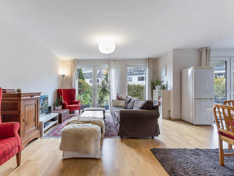 Helles Wohnzimmer mit Sofa, Sesseln, Teppich, Kühlschrank und großen Fenstern zum Garten.