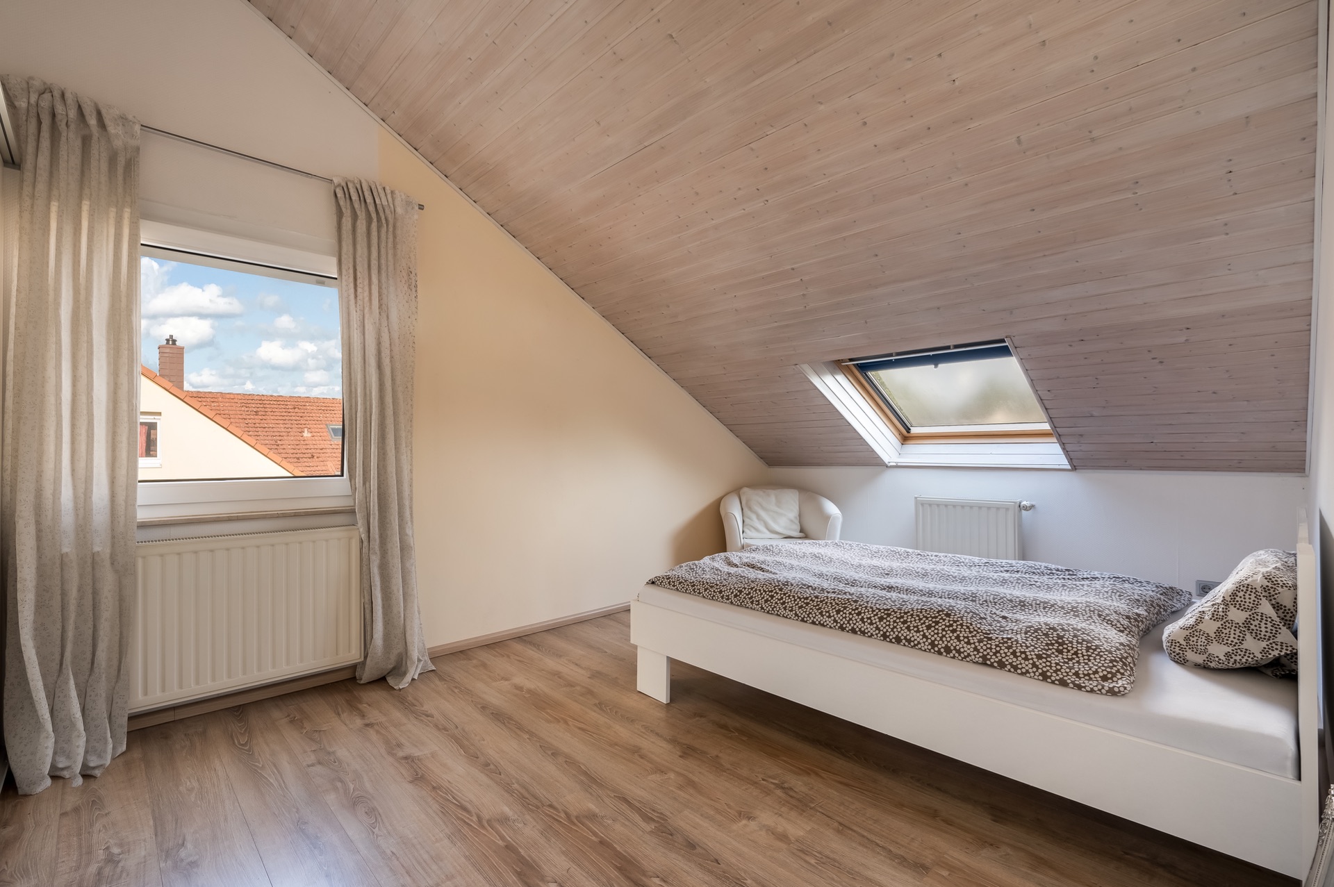 Dachzimmer mit Einzelbett, Sessel, zwei Fenstern und hellem Holzfußboden.