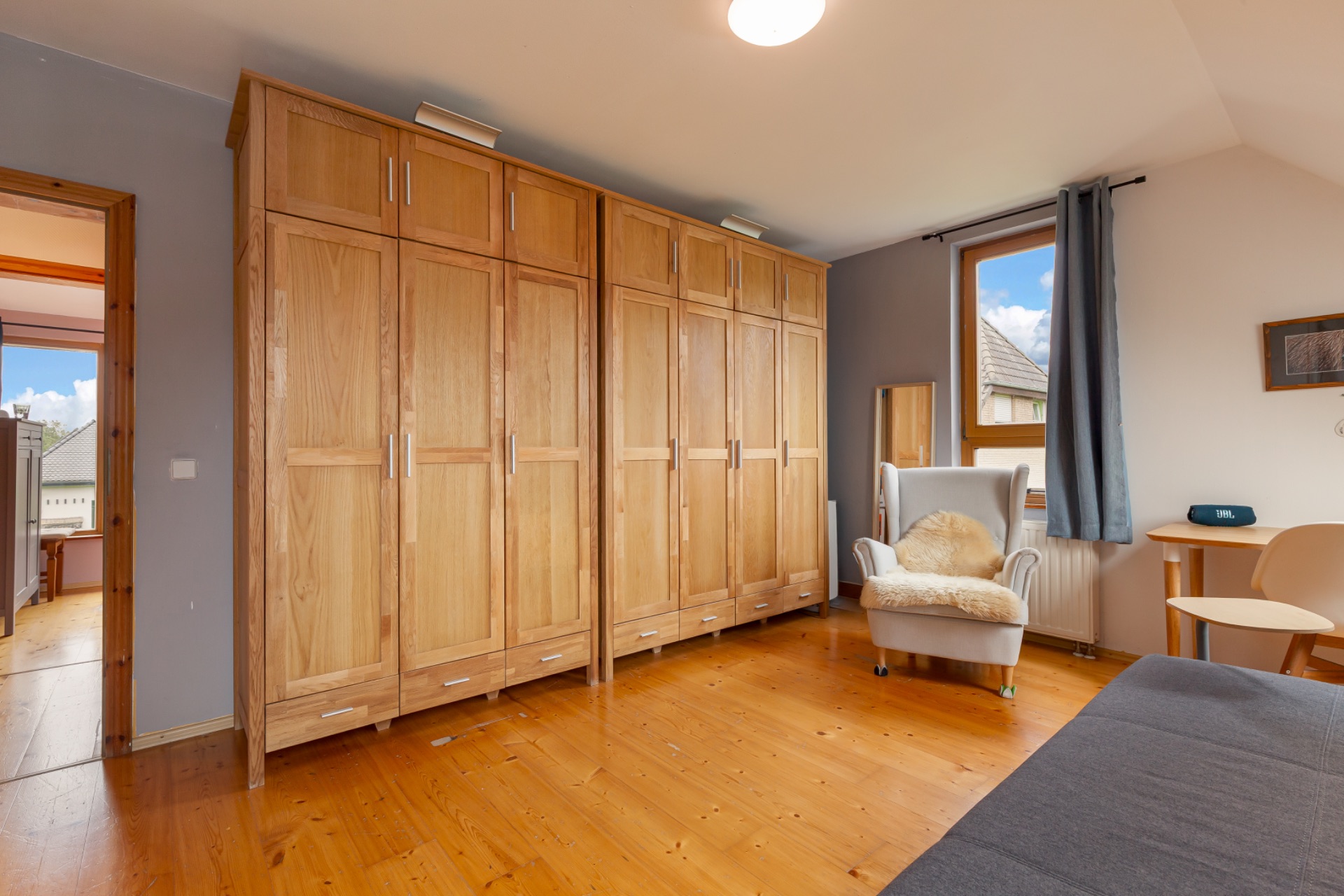 Helles Schlafzimmer mit großen Holzschränken, Sessel, Schreibtisch und Fenster mit Vorhang.