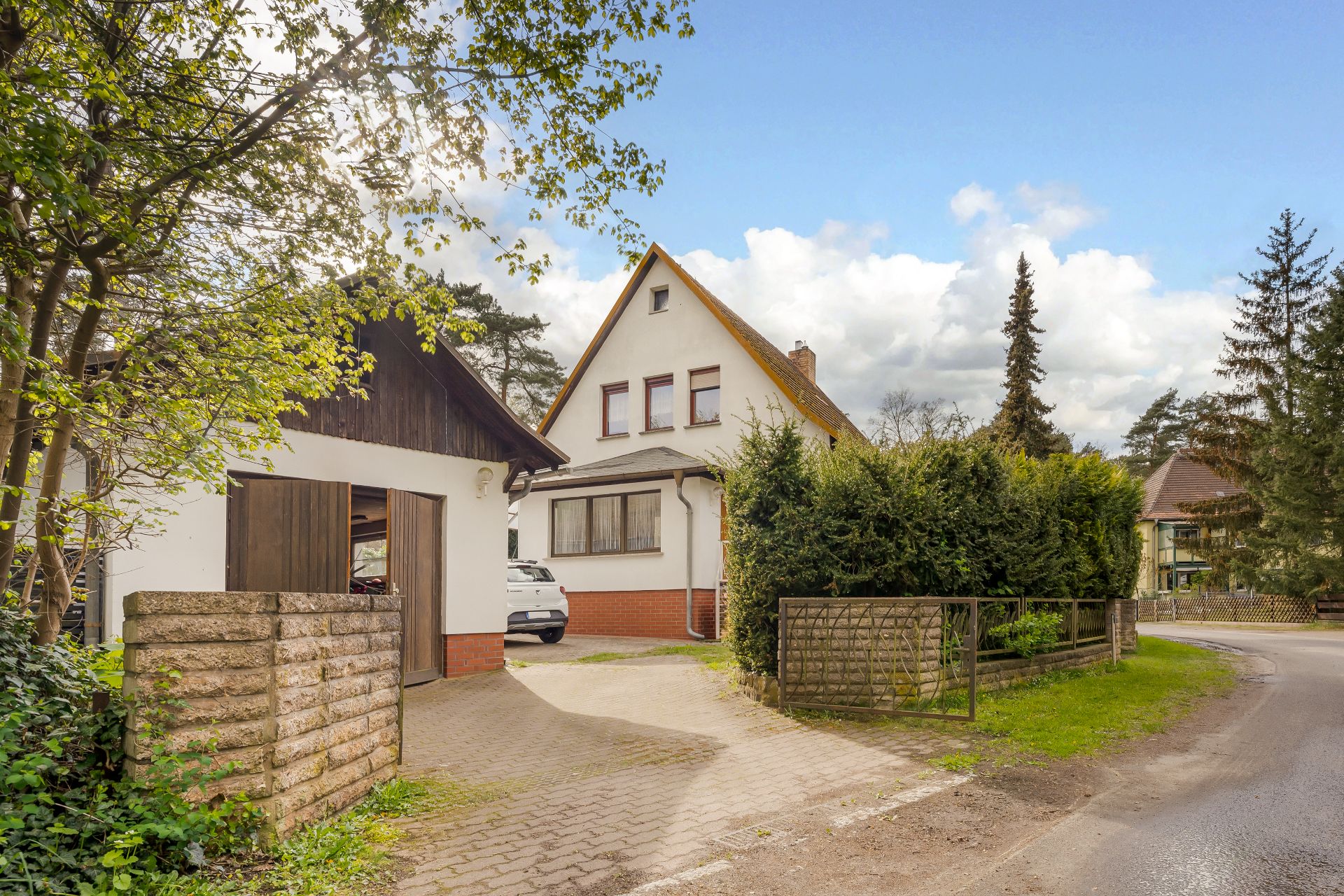 Einfamilienhaus mit spitzem Dach, Garage und Einfahrt, umgeben von Bäumen und Sträuchern.
