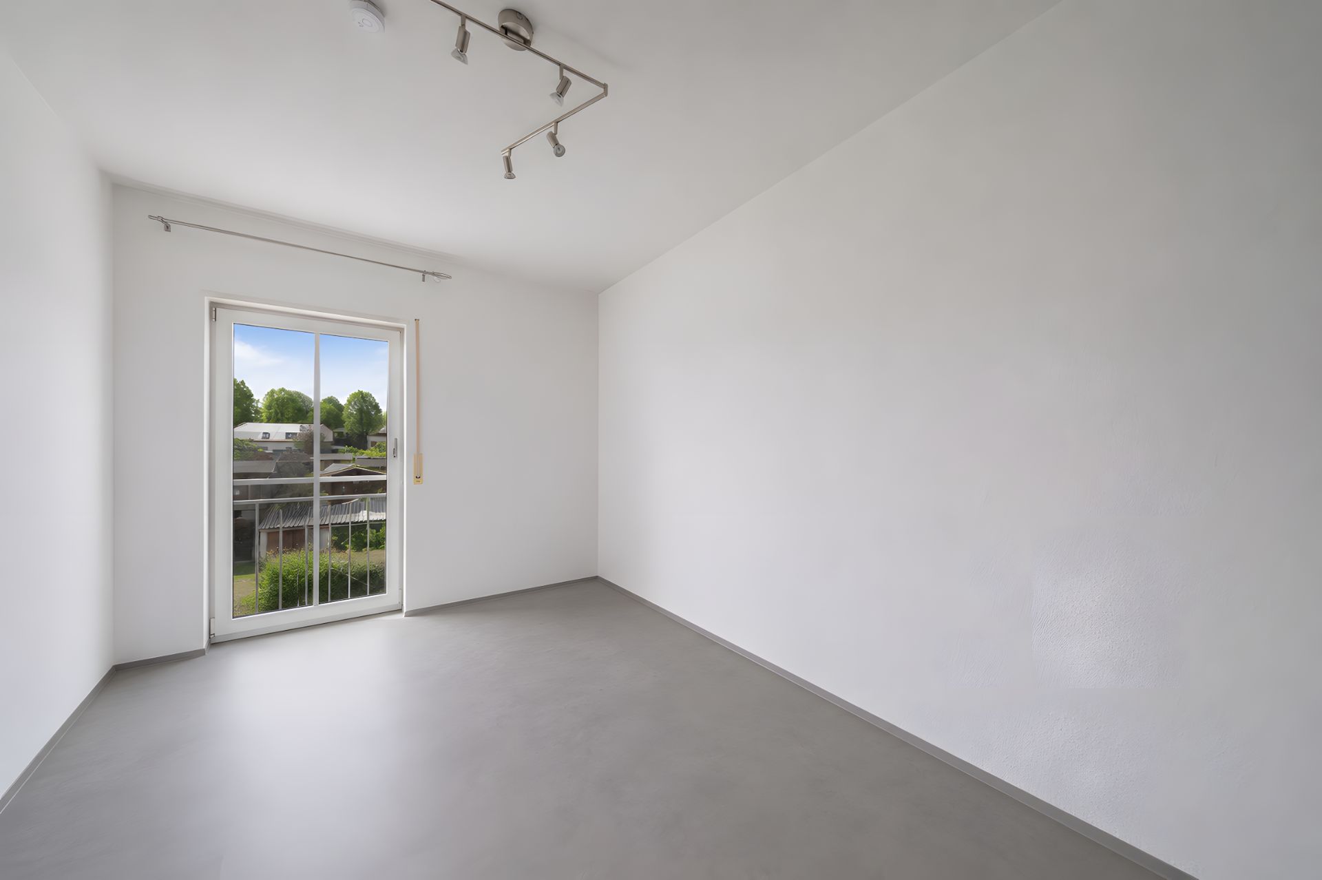 Leerer, heller Raum mit Fenster und grauem Boden, Blick nach draußen ins Grüne.