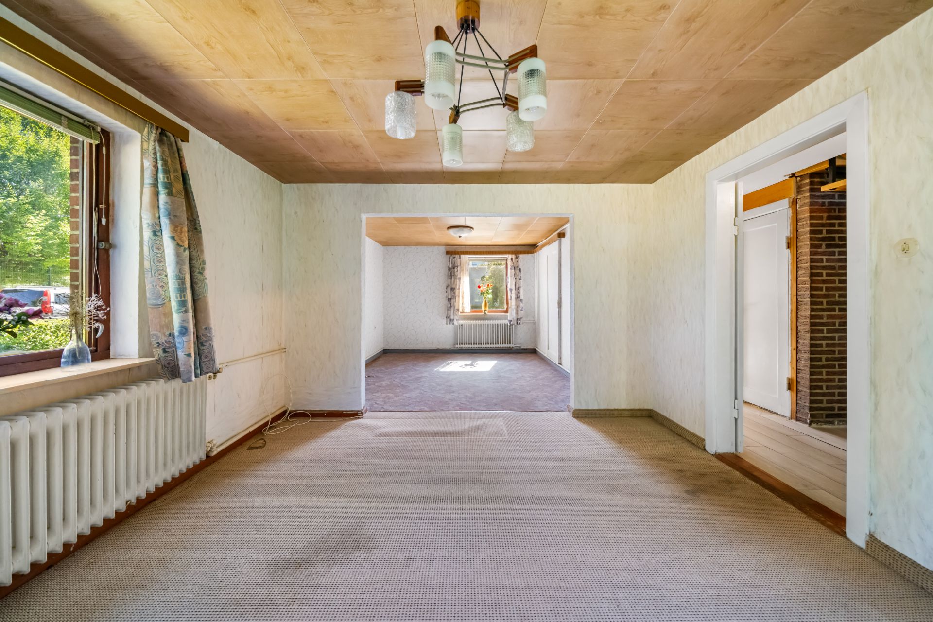 Wohnzimmer mit Teppichboden, Holzbalkendecke, Fenster, Radiator und Blick in angrenzenden Raum.