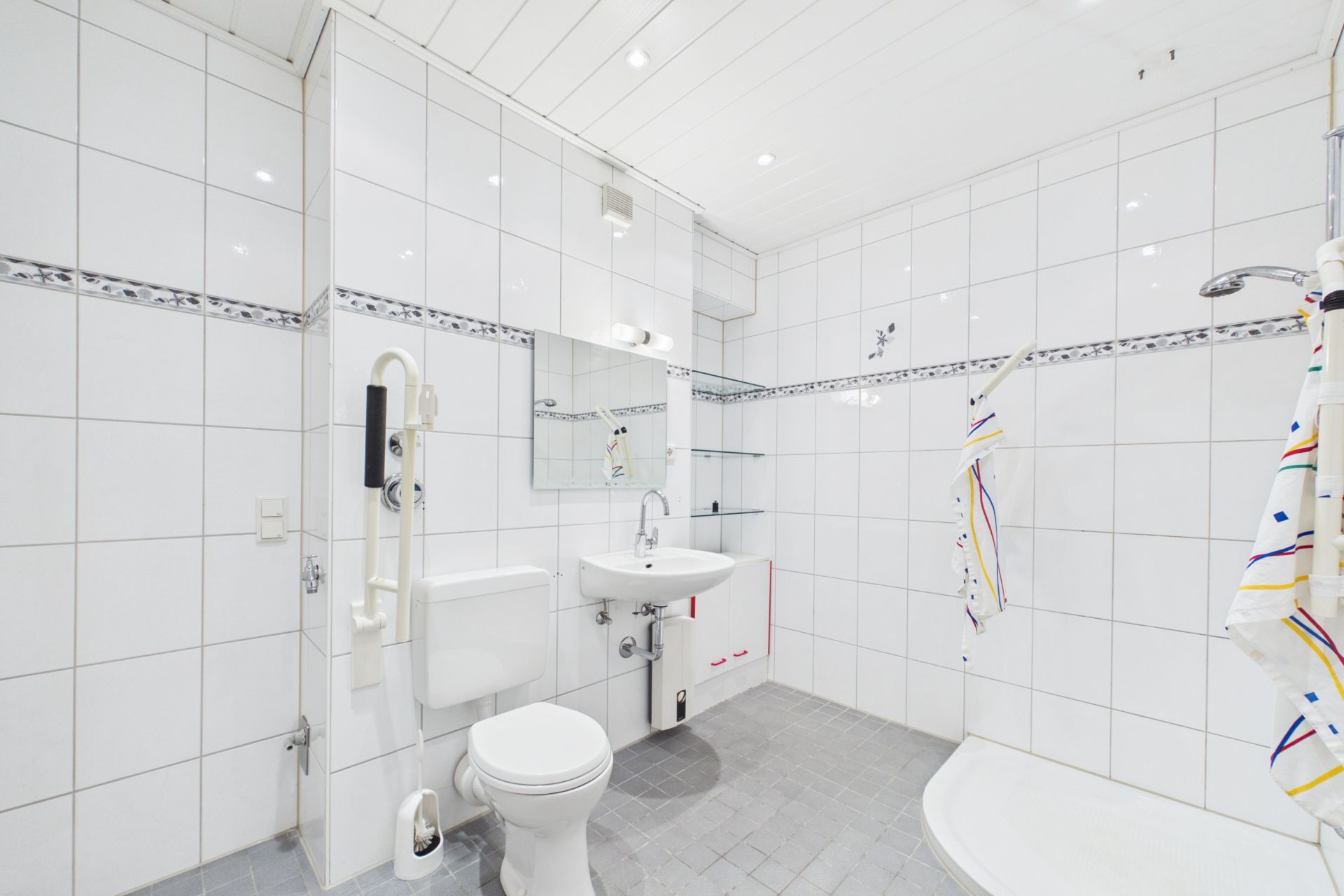 Helles Badezimmer mit weiß gefliesten Wänden, Toilette, Waschbecken und Dusche.