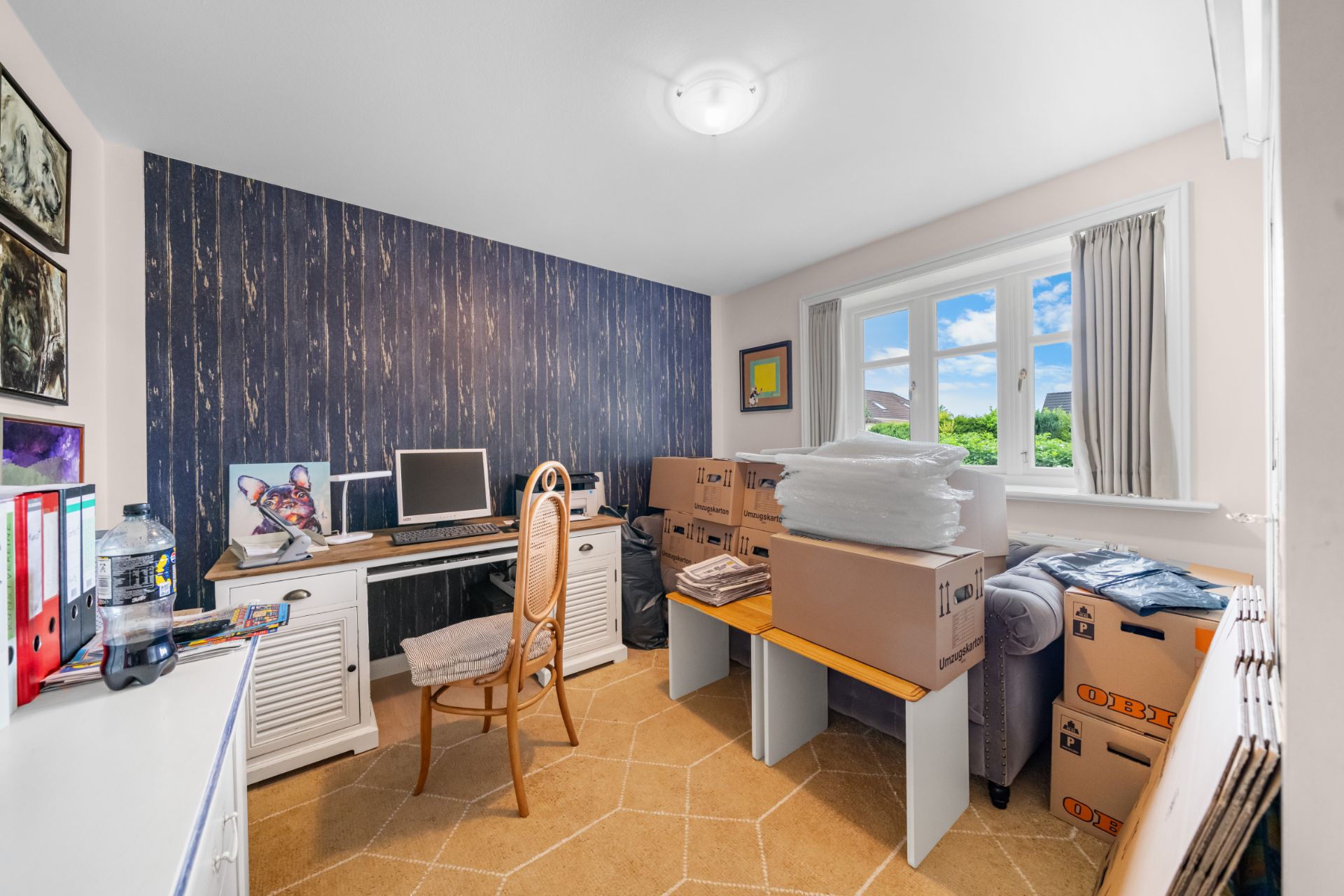 Ein Arbeitszimmer mit Schreibtisch, Computer, Stuhl, vielen Umzugskartons und großem Fenster.