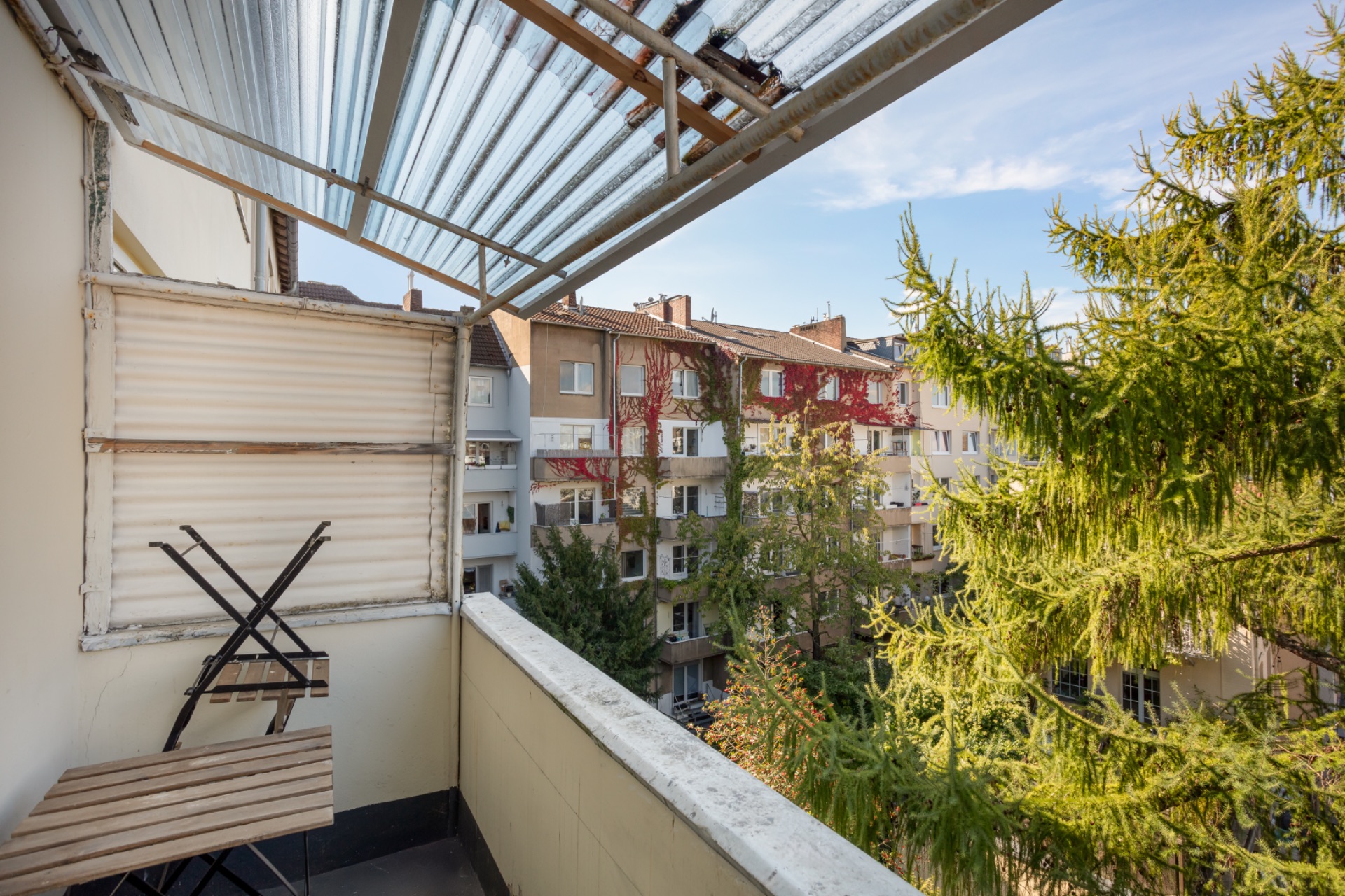 Balkon mit klappbarem Tisch und Stühlen, Blick auf grüne Bäume und Nachbarhäuser.