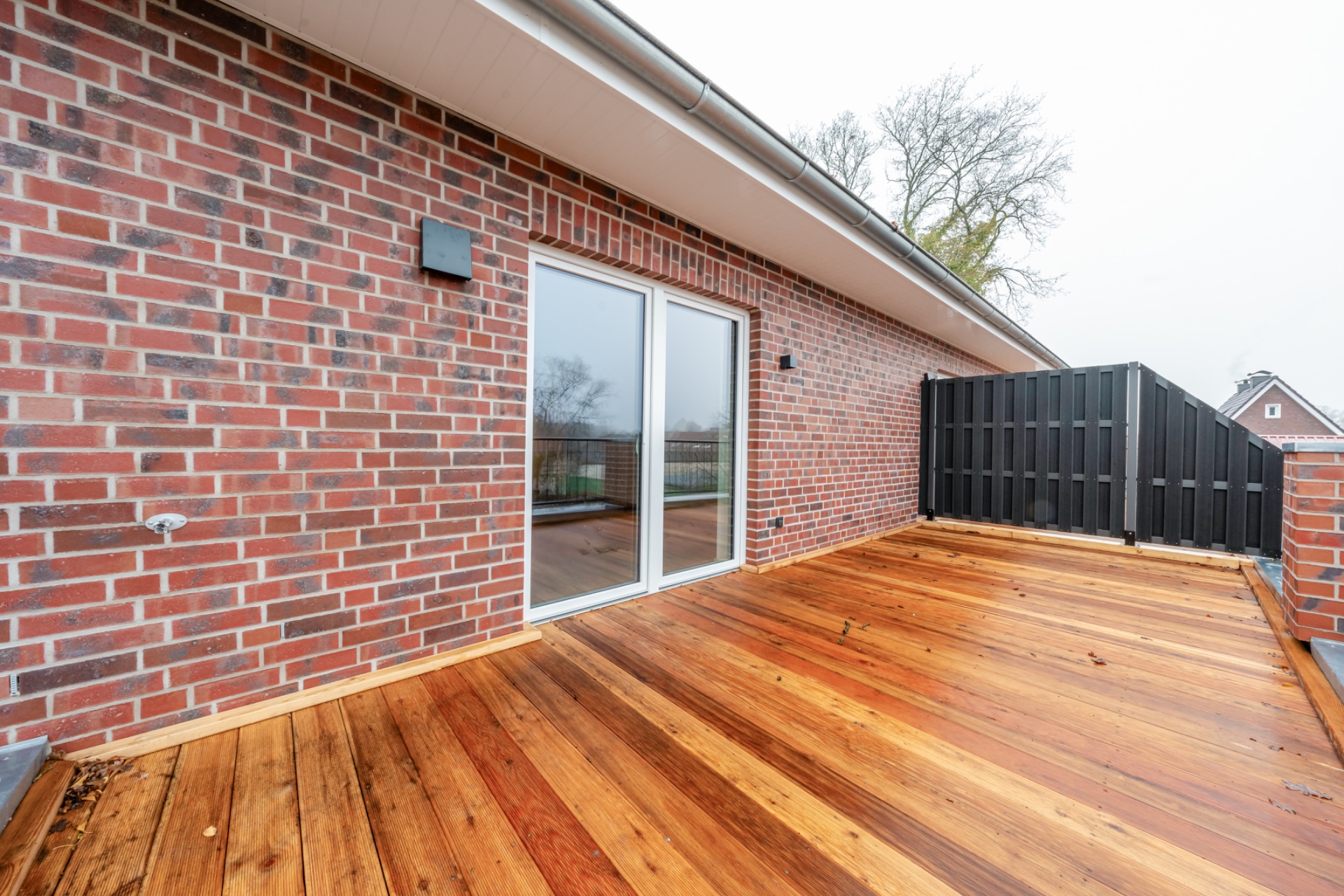 Große Terrasse mit Holzboden, Ziegelwand und Glastür, Blick auf Bäume im Hintergrund.