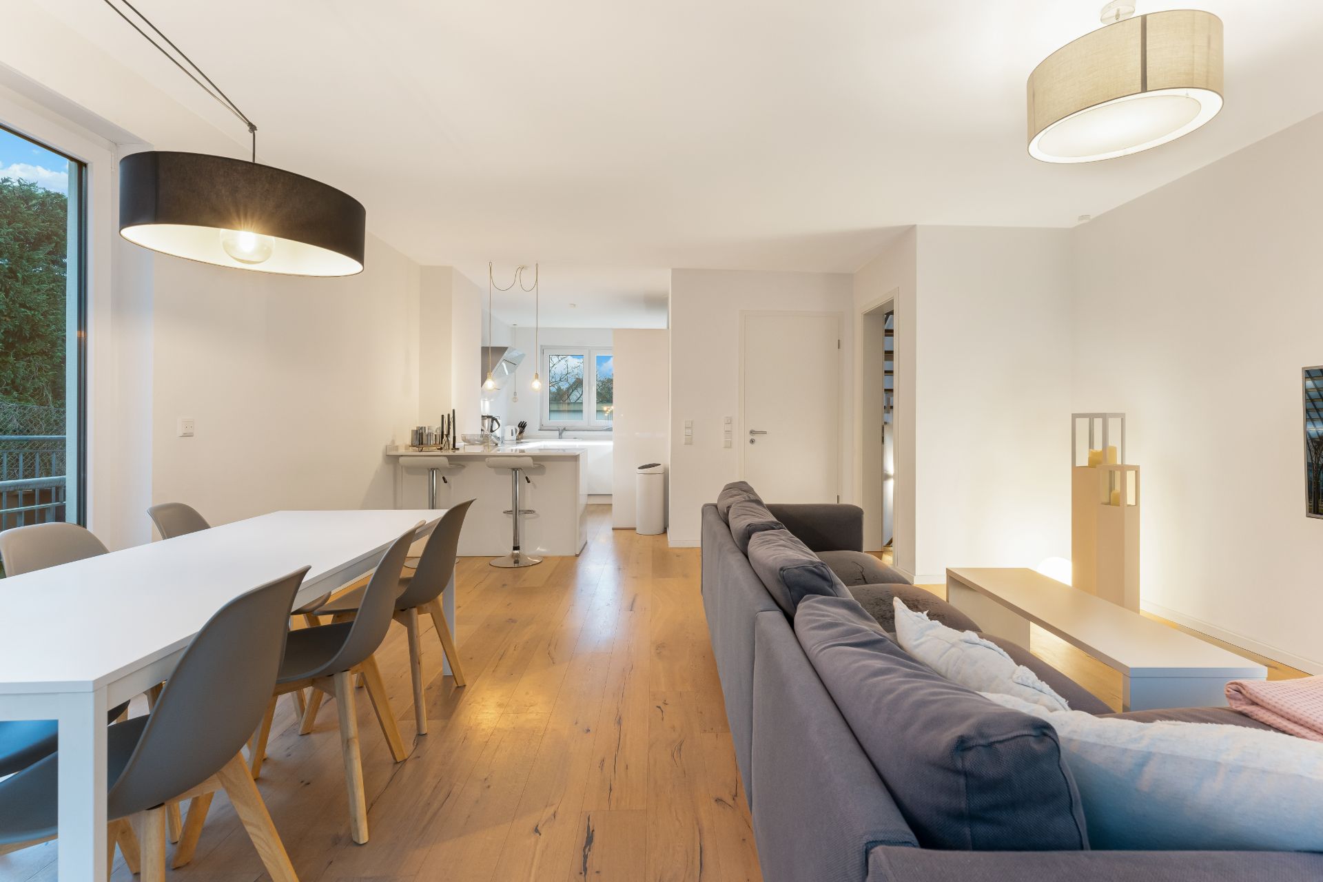 Helles Wohnzimmer mit Esstisch, Stühlen, Sofa und offener Küche, Holzboden und großen Fenstern.