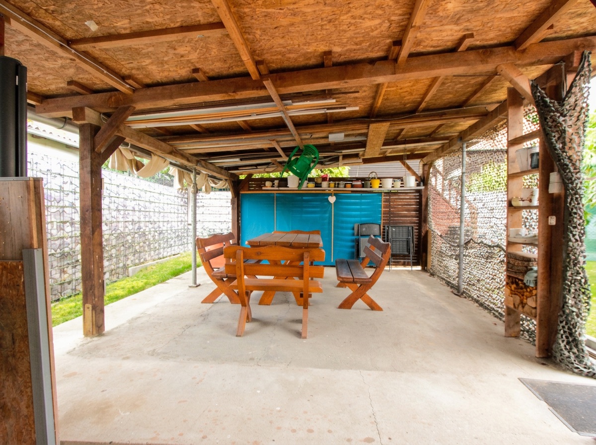 Überdachte Terrasse mit Holztisch, Bänken und Regal an der Seite.