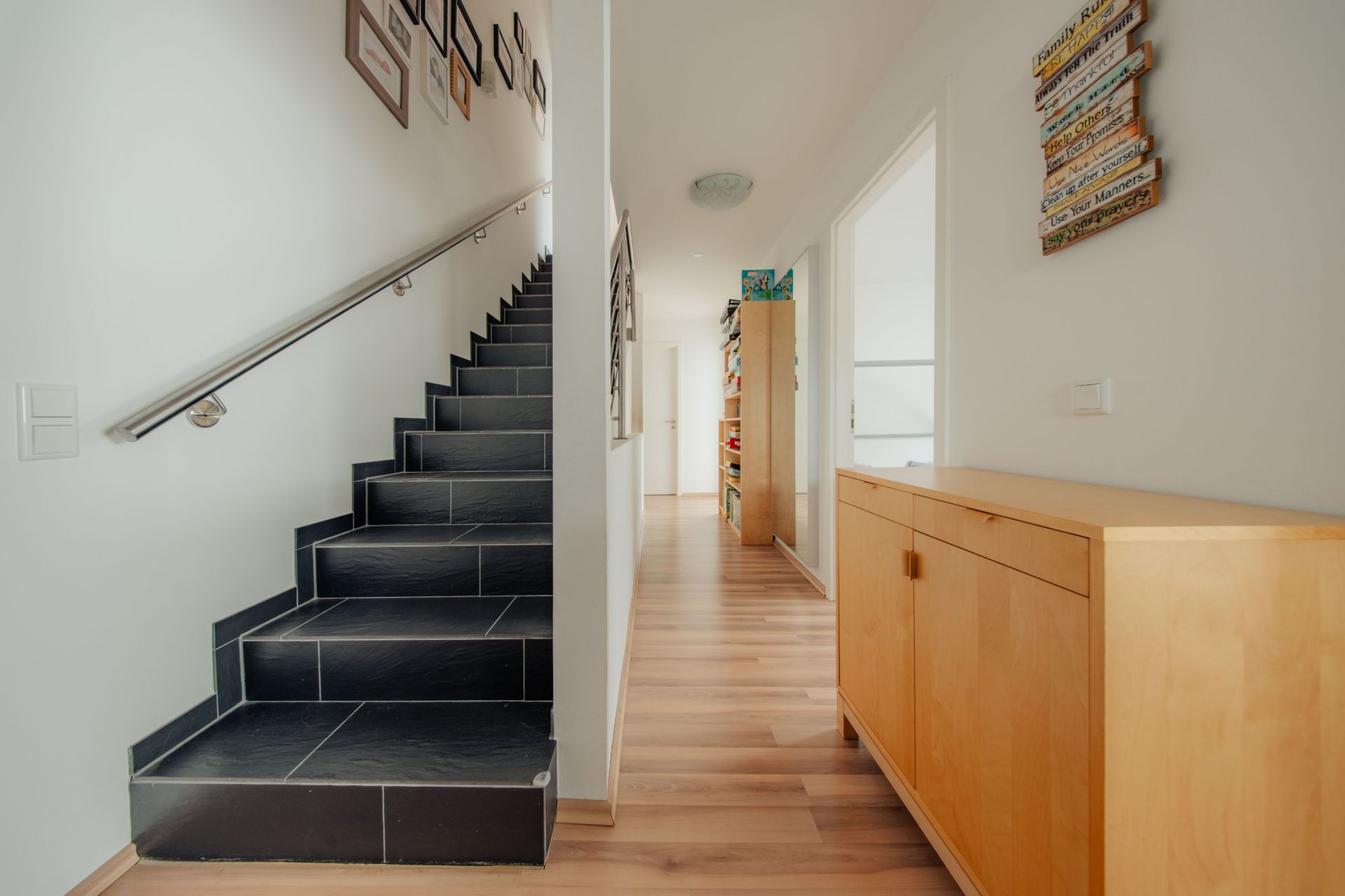 Heller Flur mit schwarzer Treppe, Holzkommode und Bildern an den Wänden.