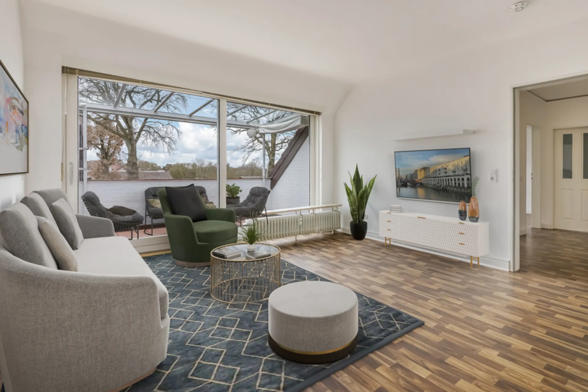 Helles Wohnzimmer mit Sofa, Sessel, Teppich, Fernseher und Zugang zu einer Terrasse mit Sitzmöbeln.
